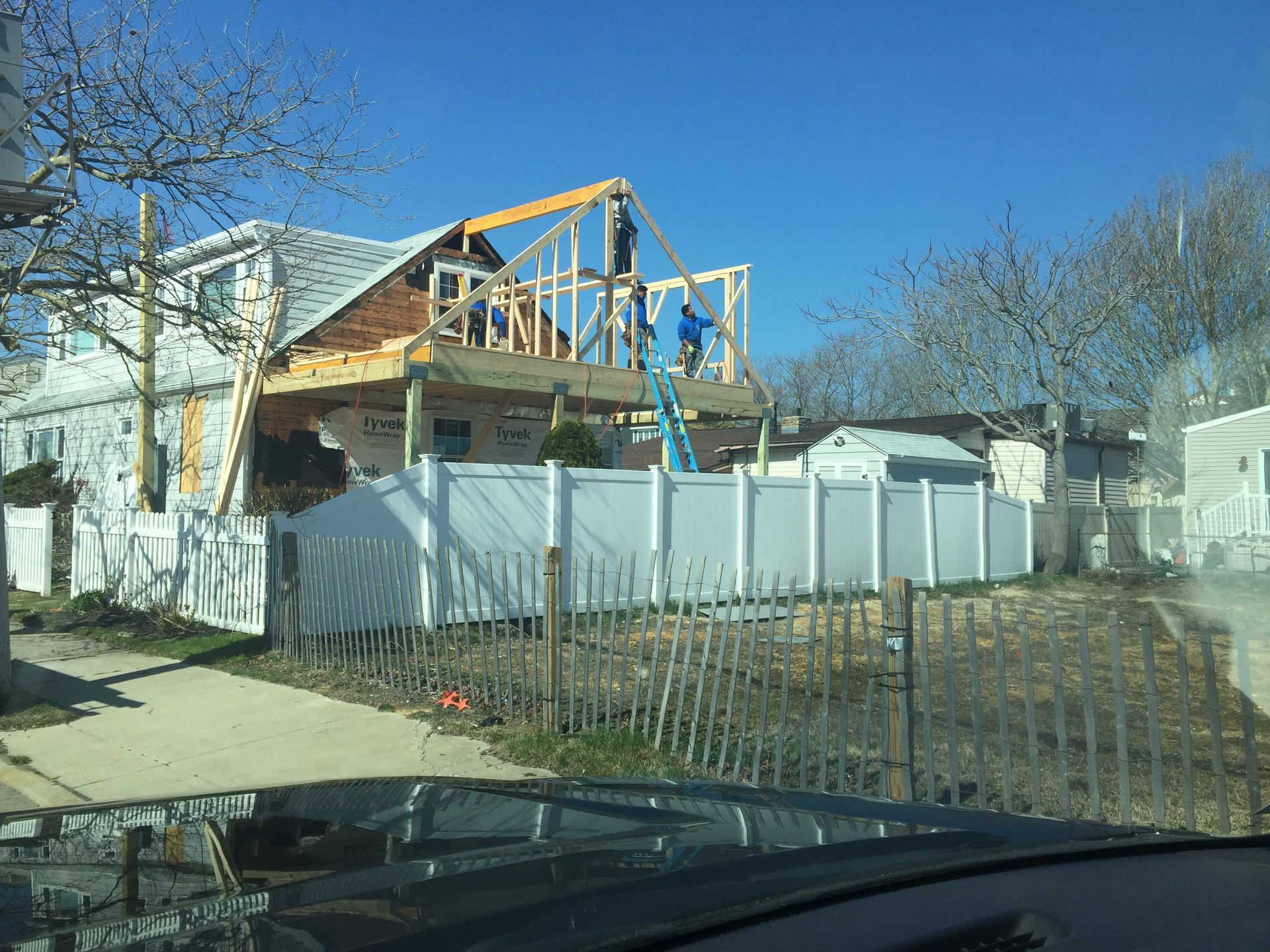 House under construction with exposed wooden framing behind a white fence on a sunny residential street