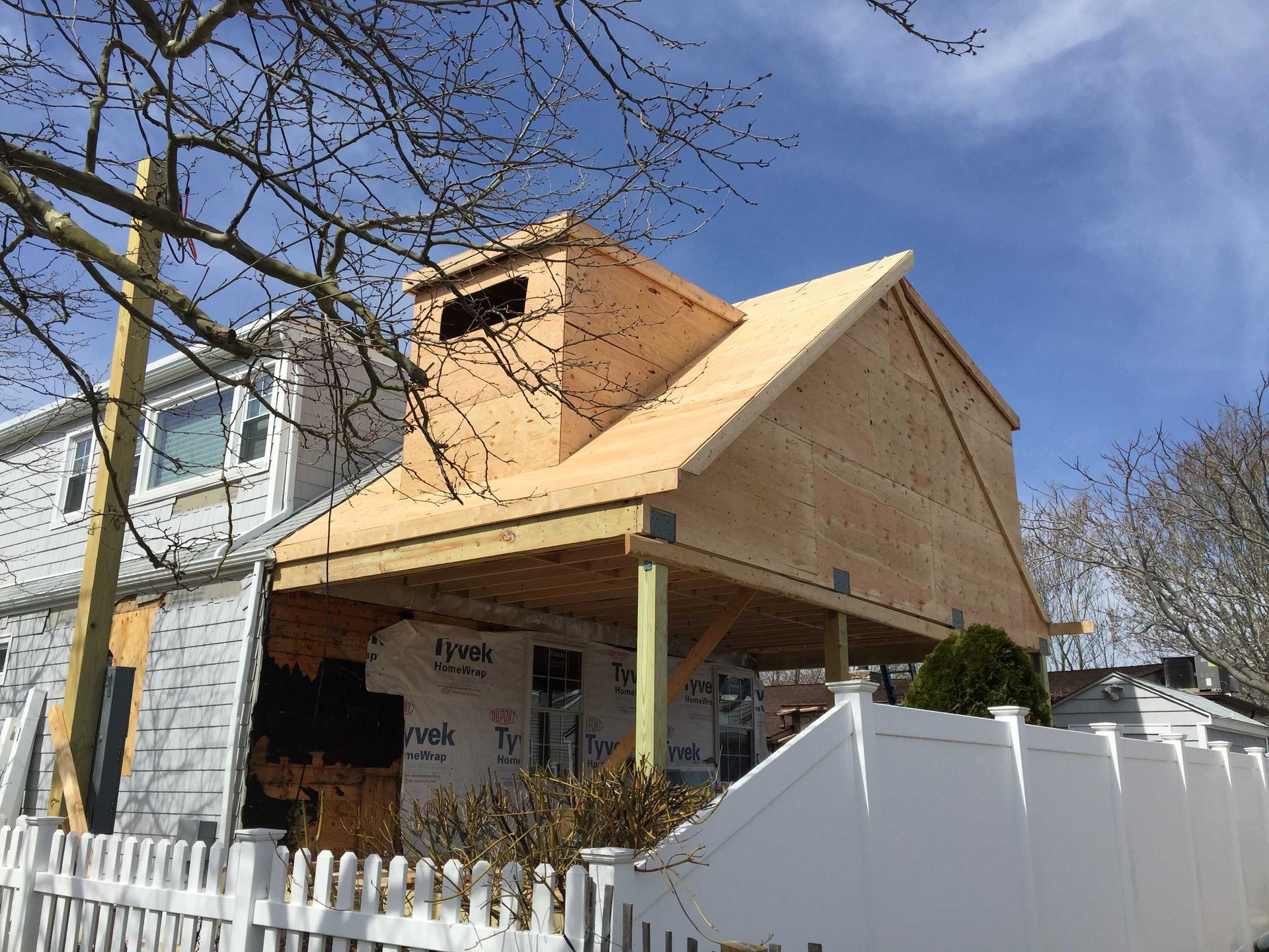 Partially built house with exposed wooden frame and white fence under a blue sky