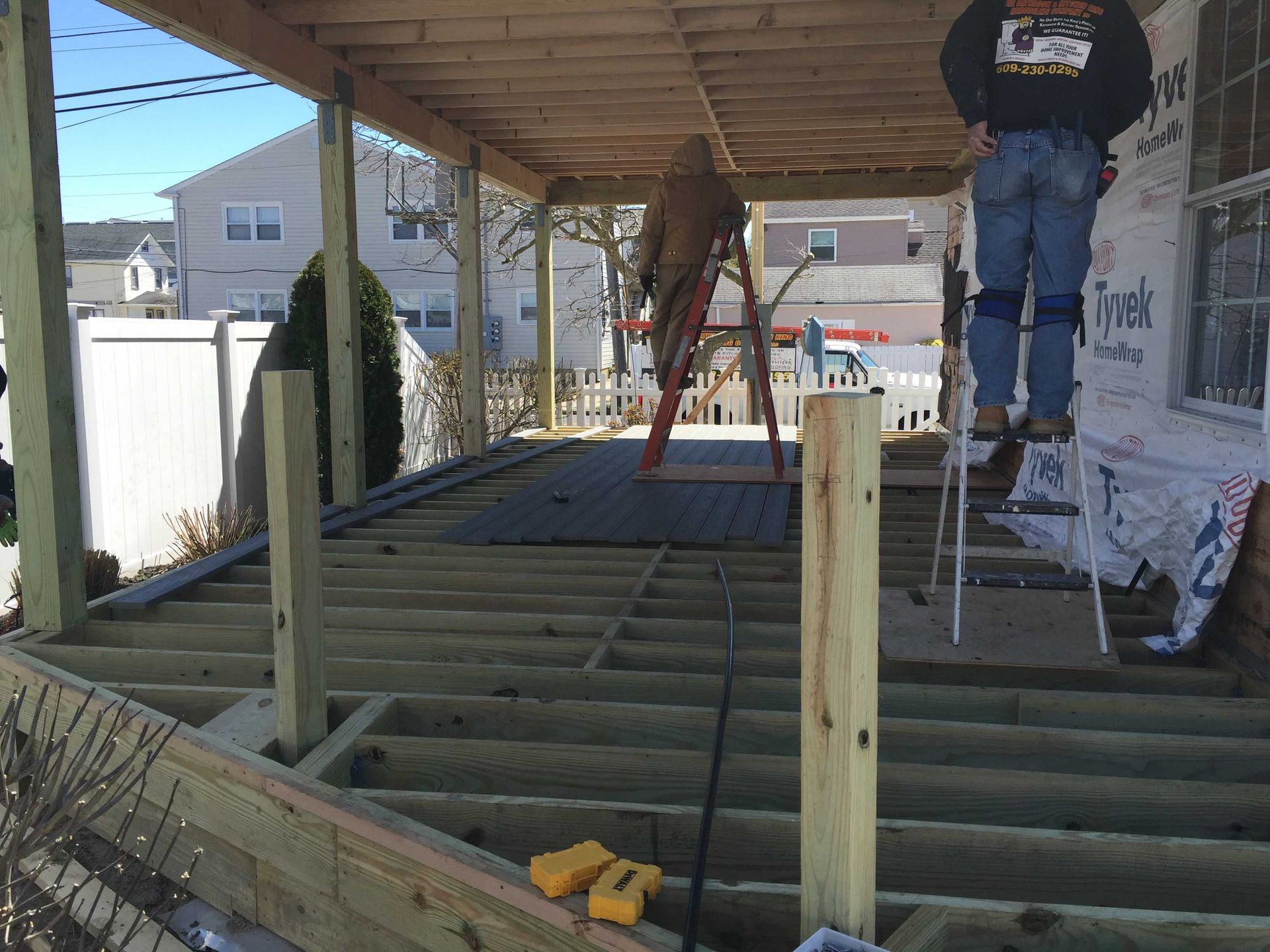 Workers framing a wooden deck under construction on a house porch, with tools and ladders nearby