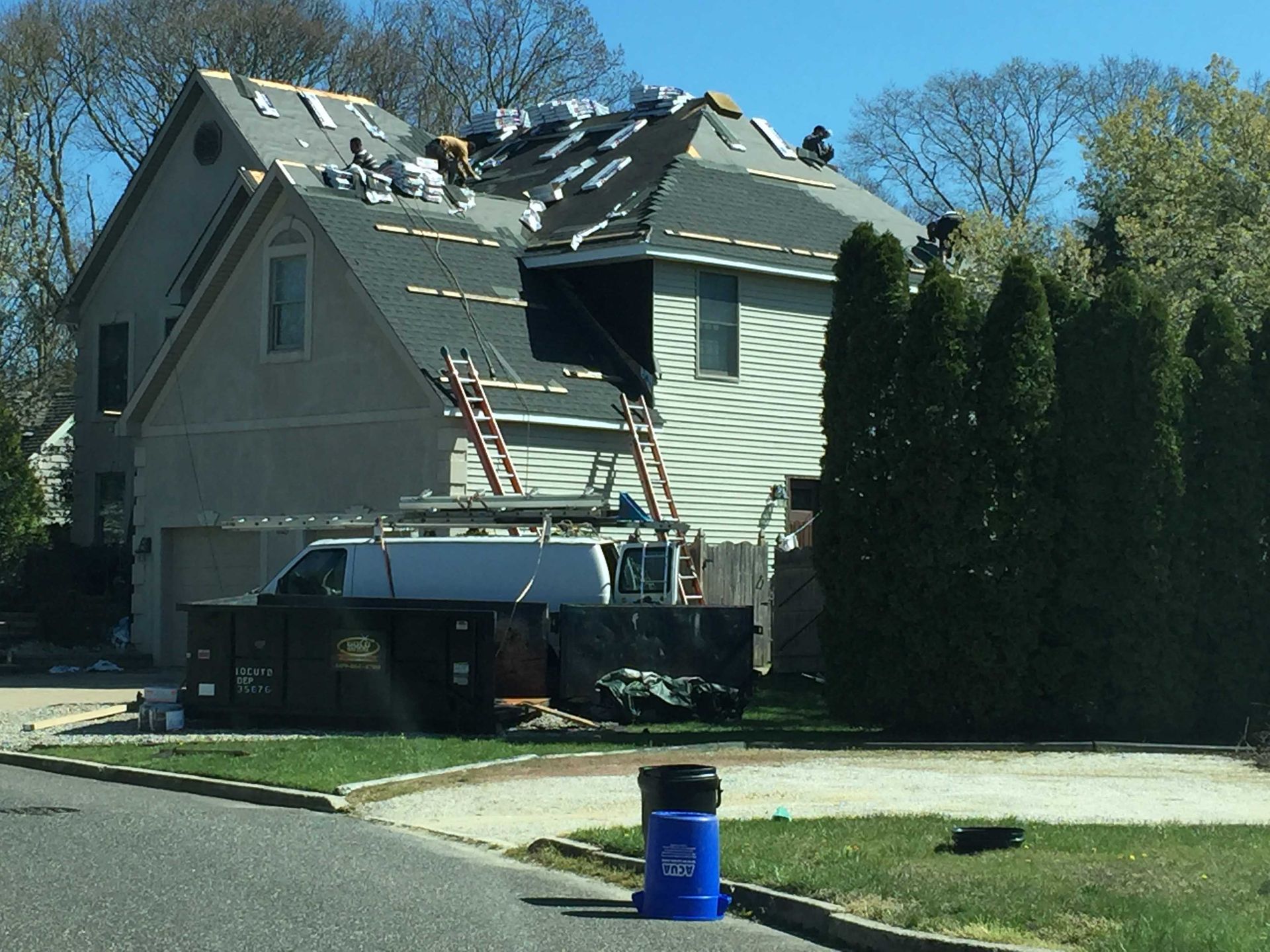 House with roof damage and ladders during repairs, next to tall hedges on a sunny street.