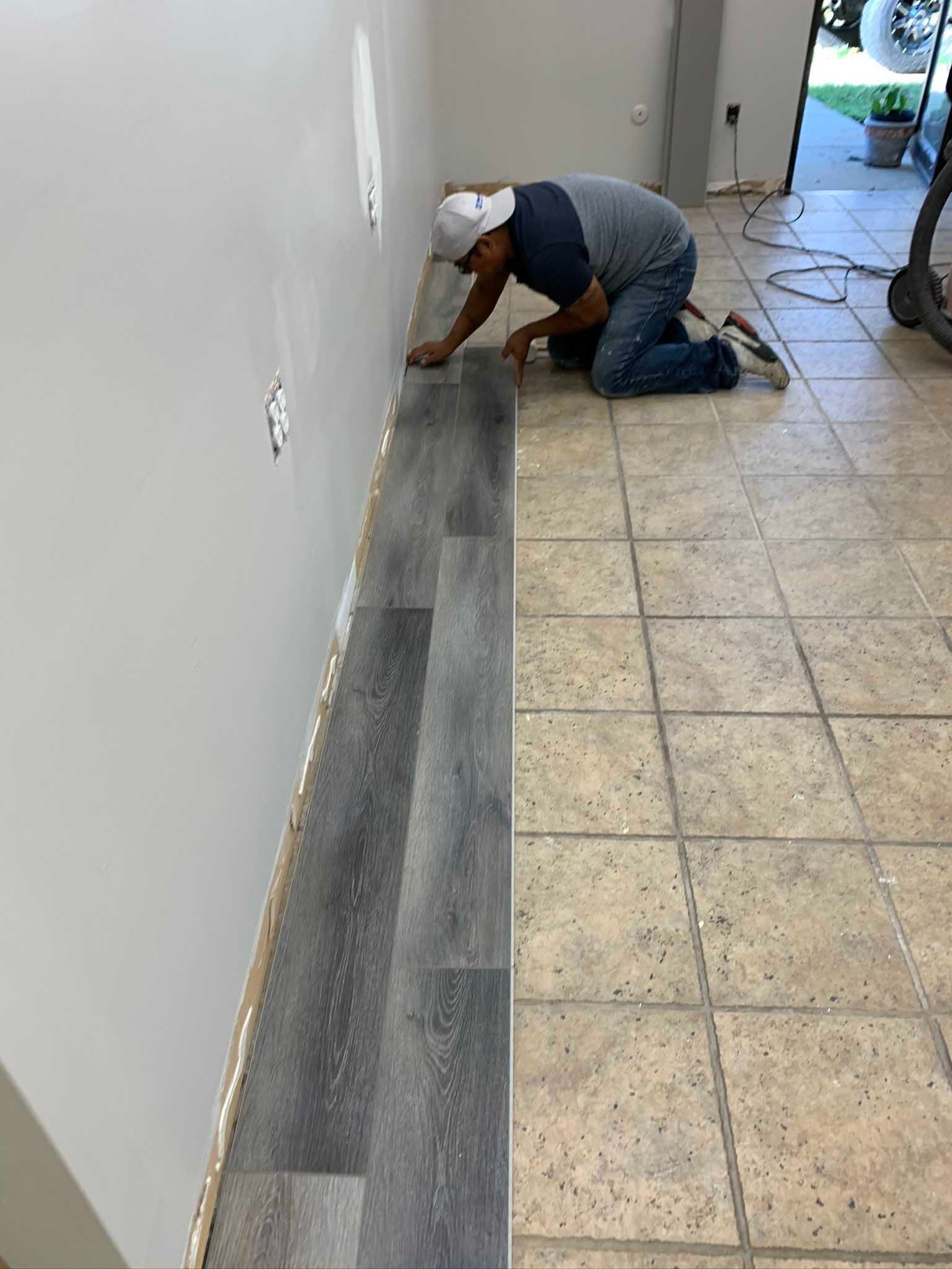 Worker kneeling by wall, installing a long gray baseboard in a tiled hallway
