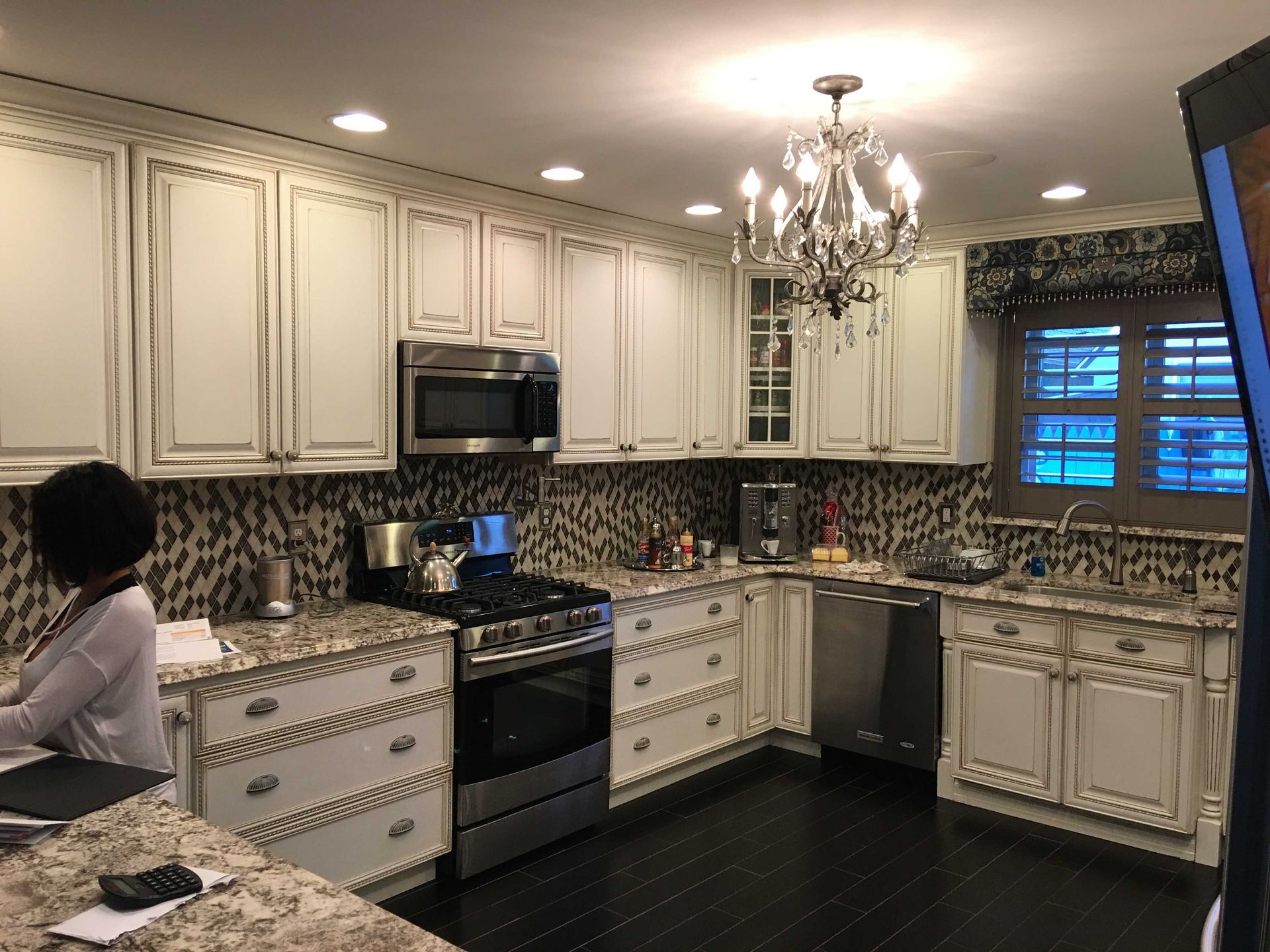 Bright kitchen with cream cabinets, mosaic backsplash, black stove, chandelier, and a person at the counter.