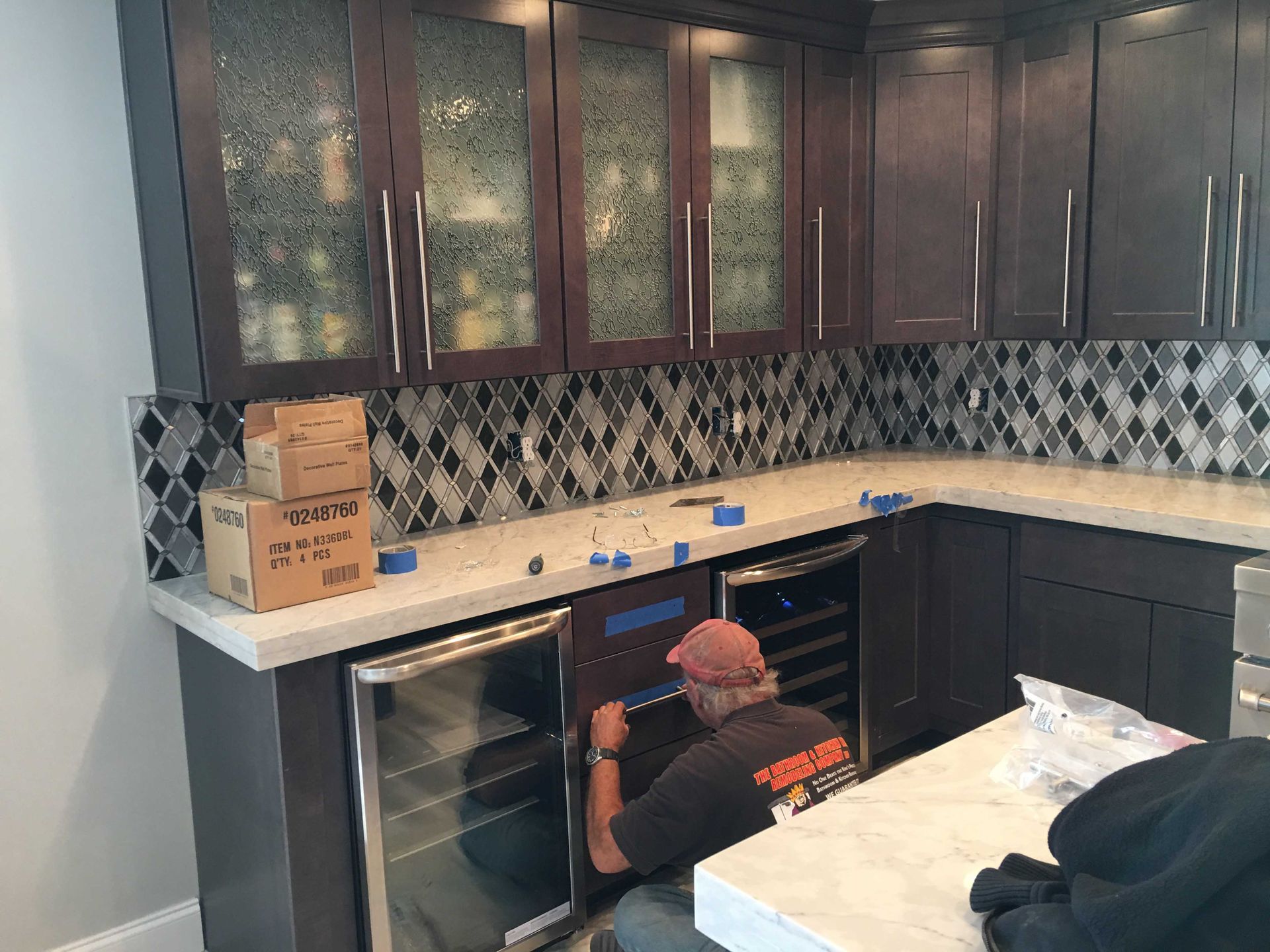 Person crouching in a kitchen under cabinets, working near a sink and countertop with boxes and supplies nearby