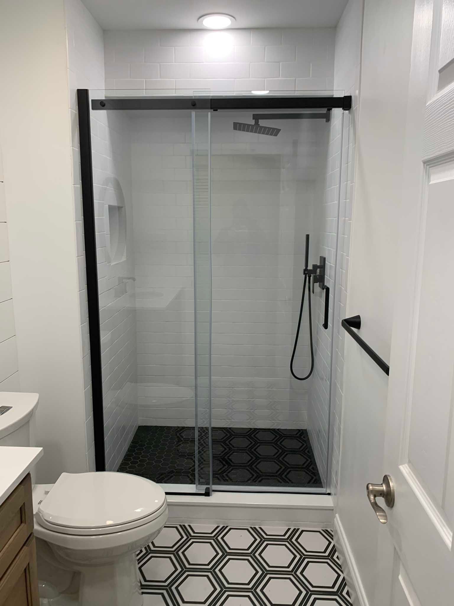 Small bathroom with a glass shower, white walls, black fixtures, and patterned floor tiles.