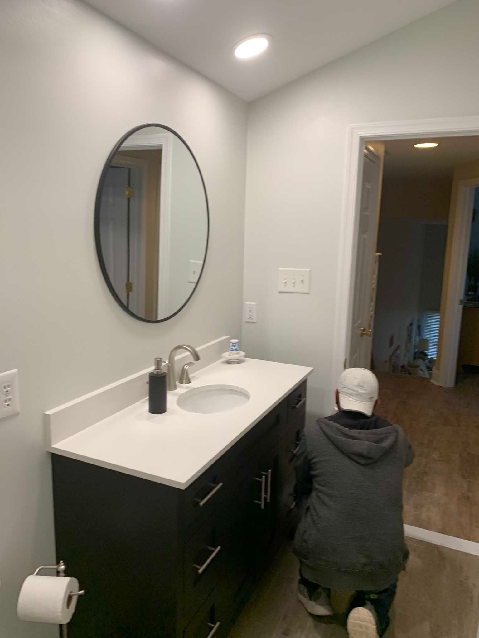 Person kneels by a bathroom vanity with an oval mirror and white countertop in a gray room.