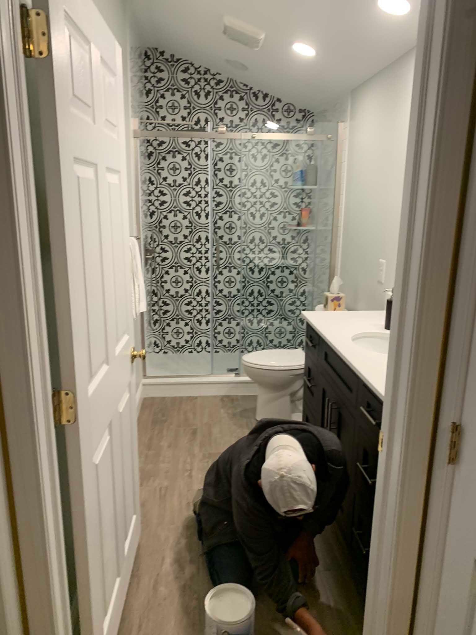 Person kneeling on bathroom floor while installing or cleaning near a vanity and shower with patterned tile.