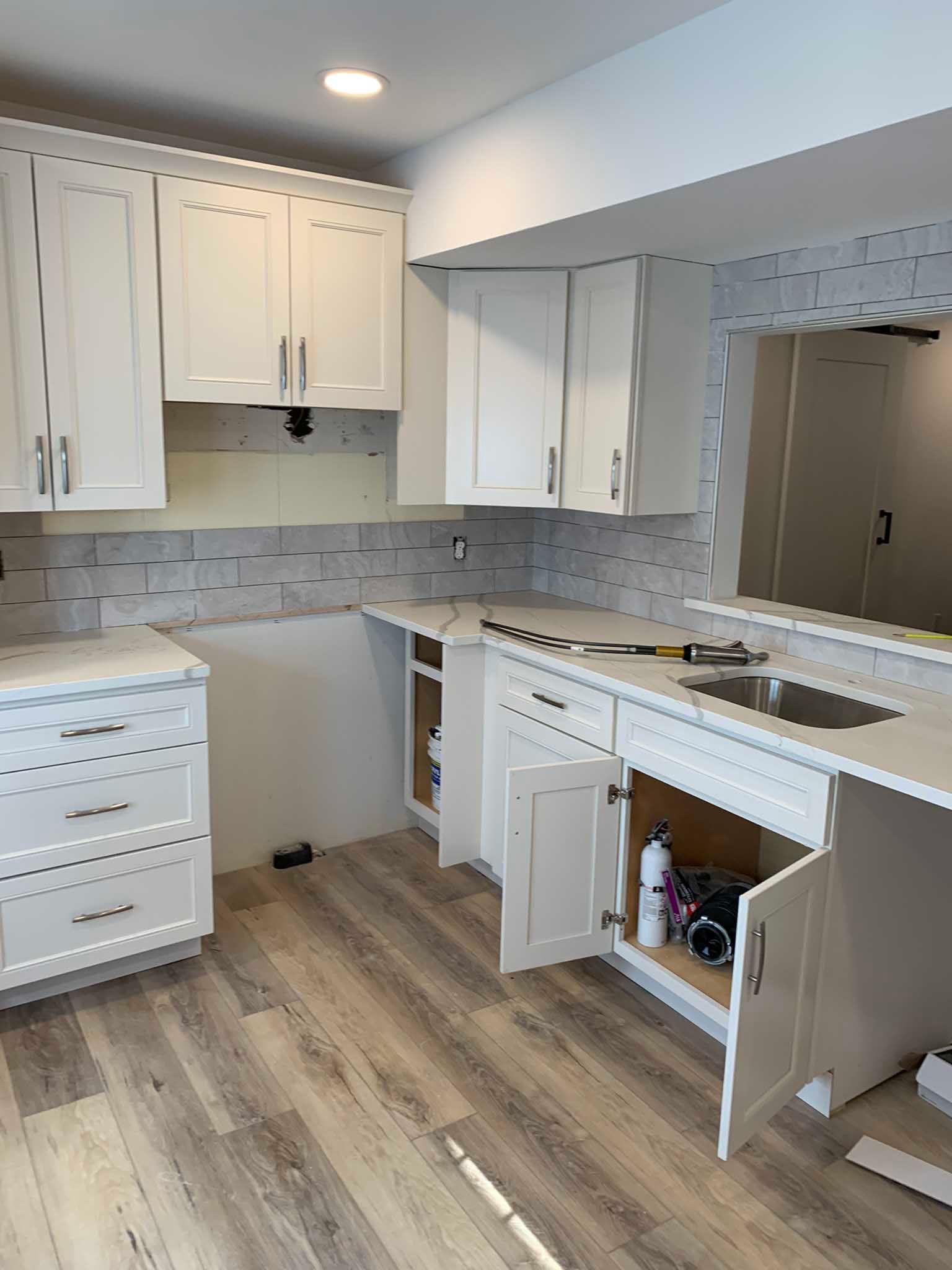 Bright white kitchen with open cabinets, tiled backsplash, wood floor, and a sink with cabinet doors open.