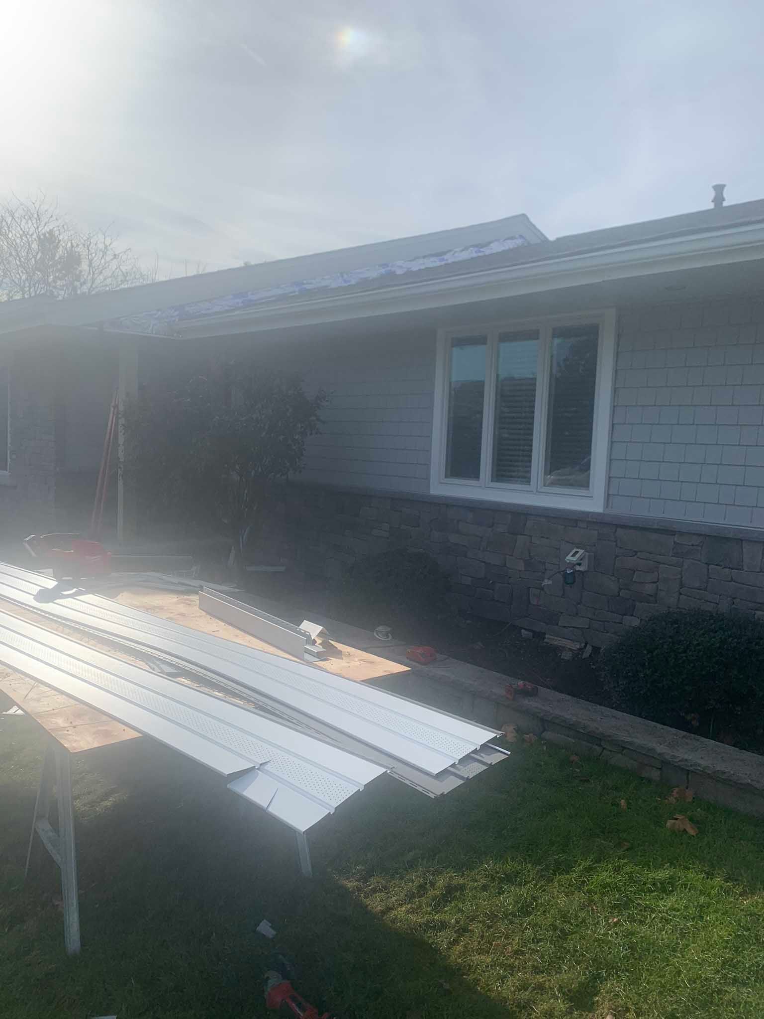 House exterior with stacked white metal roofing panels in the yard, sunlight glare on the left