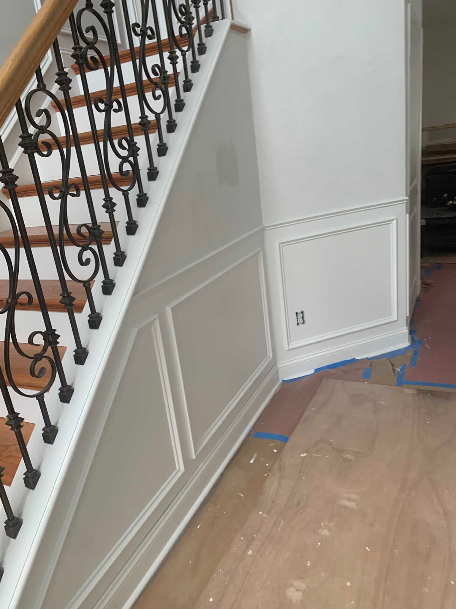 Staircase with ornate iron railing and white wainscoting; floor is covered with protective plastic during renovation.