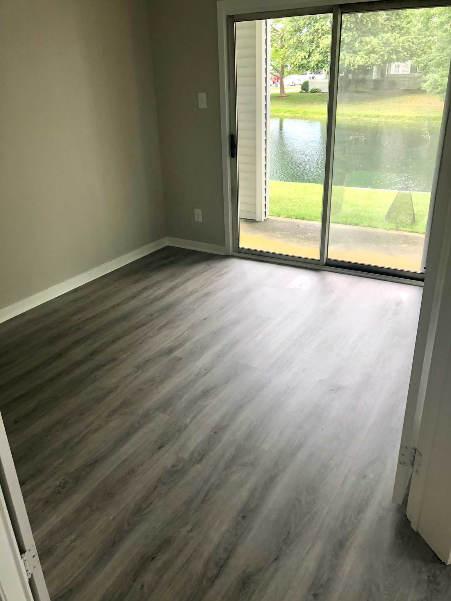Empty room with gray wood flooring and sliding glass door overlooking water and greenery
