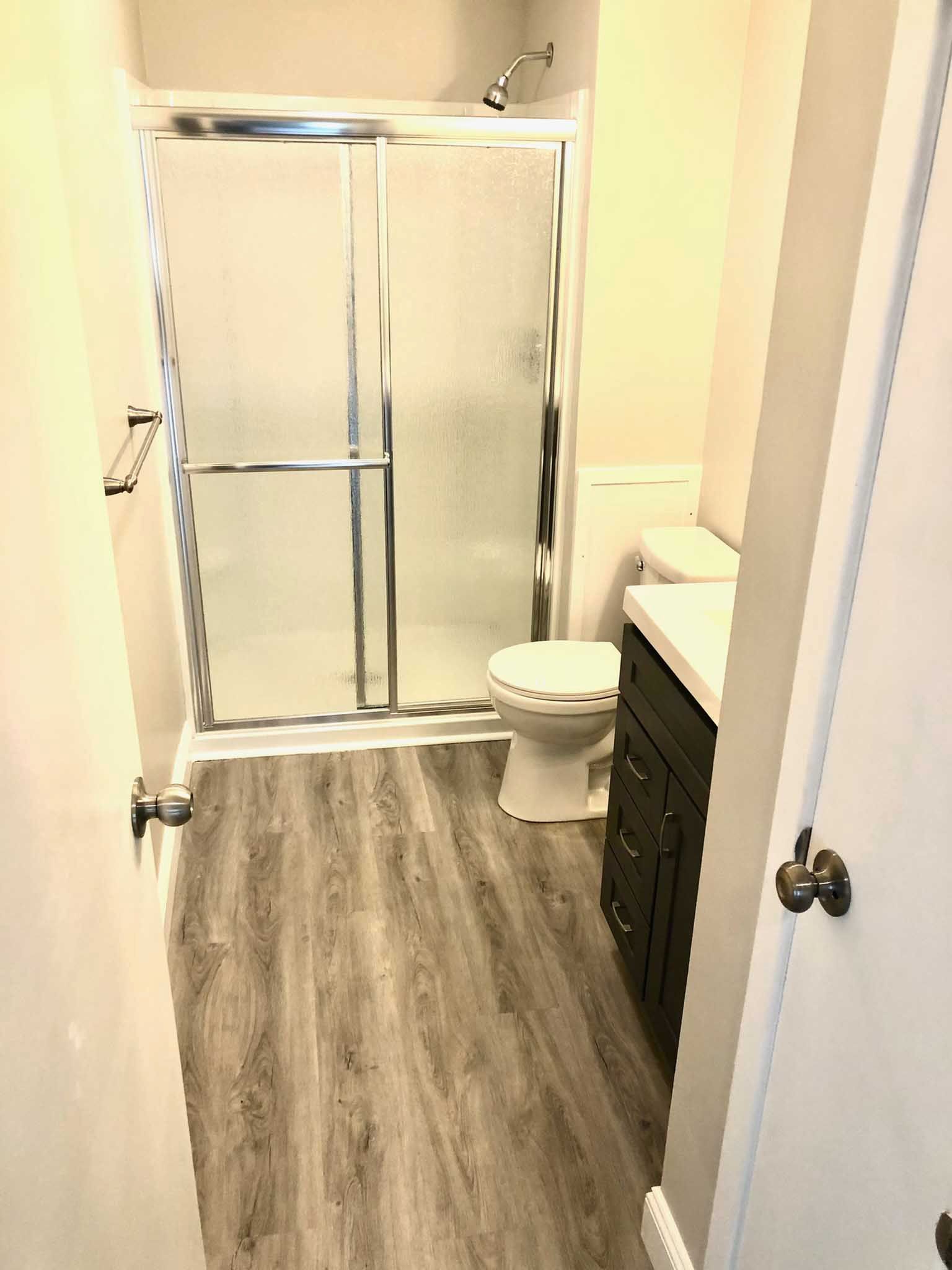 Small bathroom with shower stall, toilet, sink vanity, and gray wood-look floor.