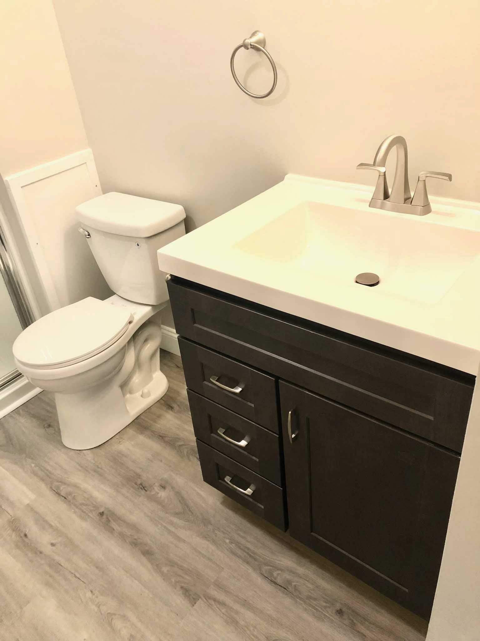 Small bathroom with white toilet and sink vanity with dark cabinets and gray wood flooring.