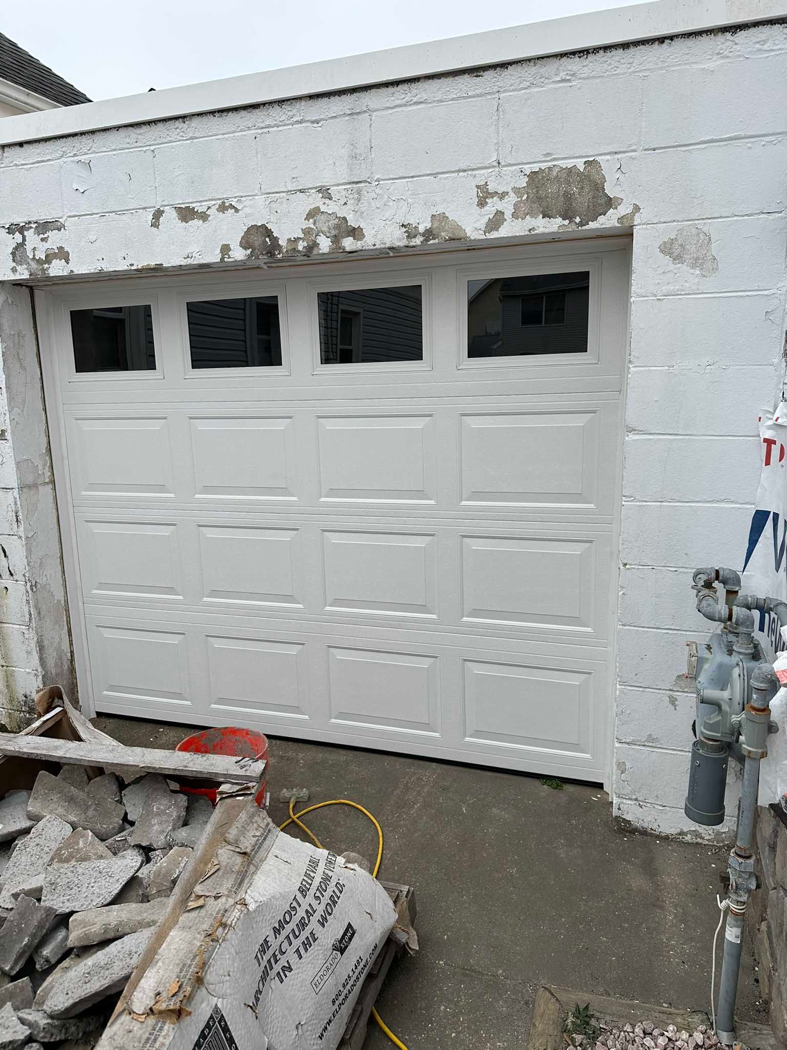 Partially installed white garage door in a construction area with debris and exposed utility pipes