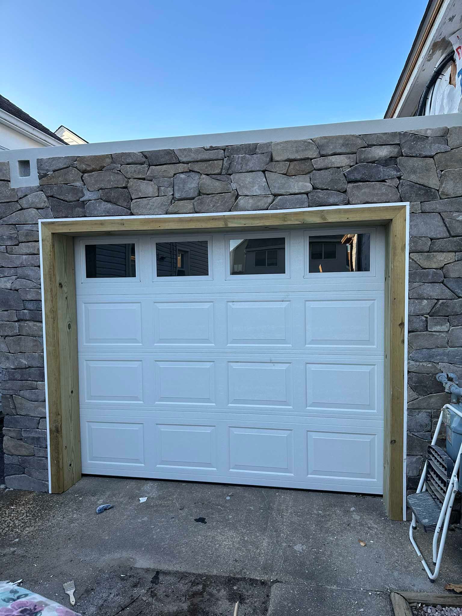 White garage door framed in beige trim set into a stone wall under a blue sky