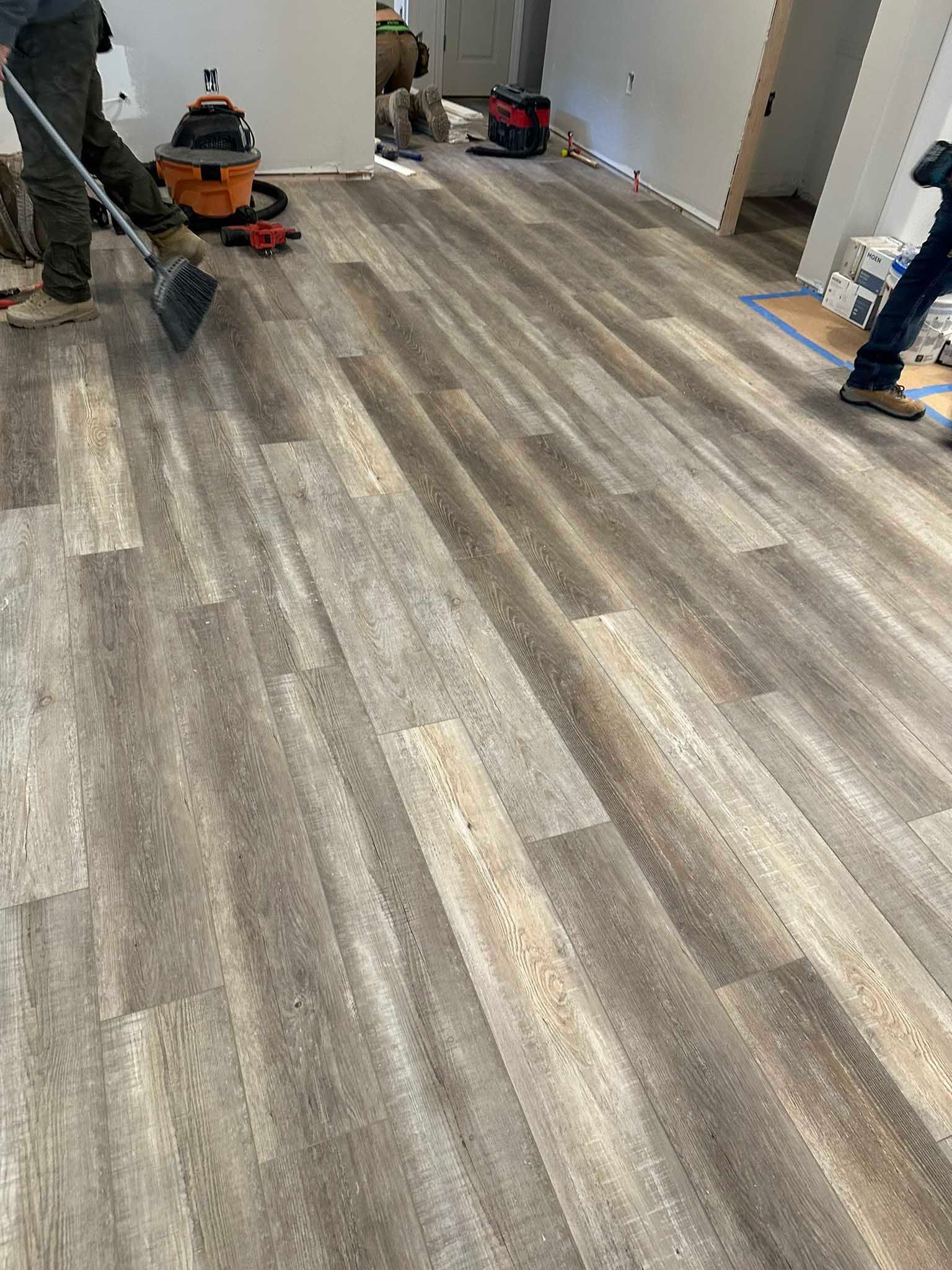 Workers installing gray wood-look flooring in a hallway, with tools and supplies along the walls.