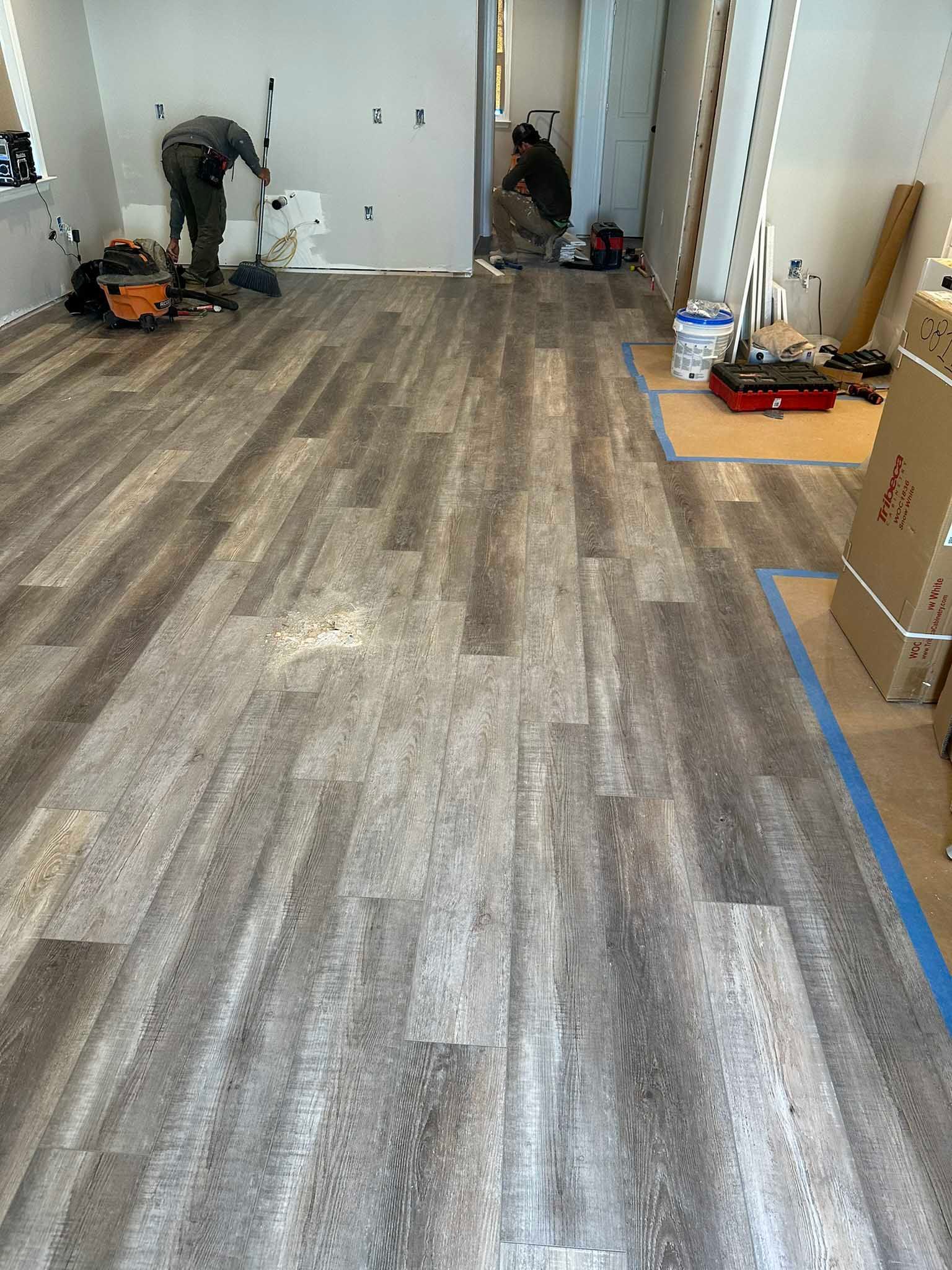 Worker installing wood-look flooring in a long room with tools and materials along the walls