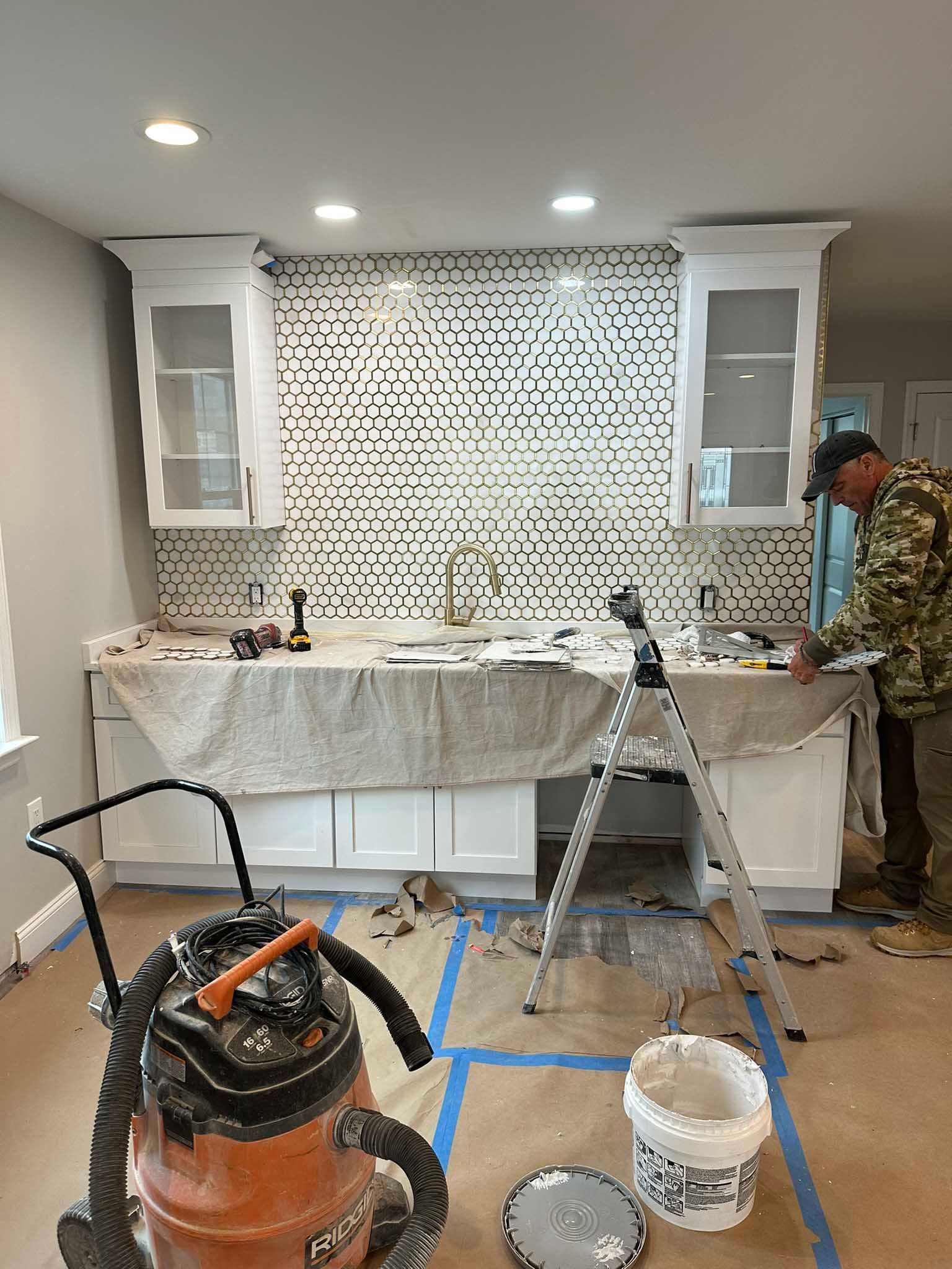 Kitchen under renovation with tiled backsplash, sink cabinets, tools, and a worker at right