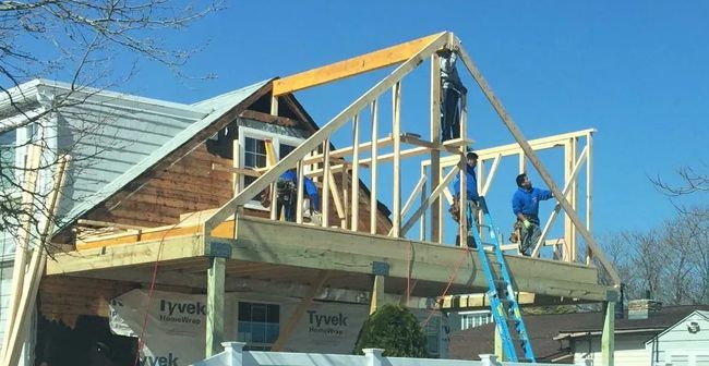 Workers framing a house roof with wooden beams, one on a blue ladder under a clear sky.