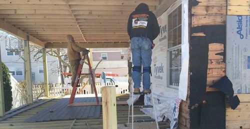 Two workers repair siding on a house porch, one on a ladder and one standing on scaffolding.