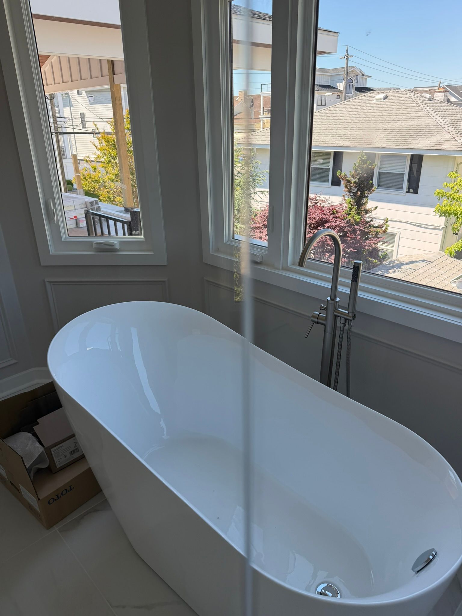 White freestanding bathtub by large windows overlooking a sunny neighborhood street