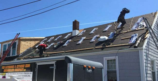 Two workers on a gray roof repairing shingles beside a black trailer under a clear blue sky.