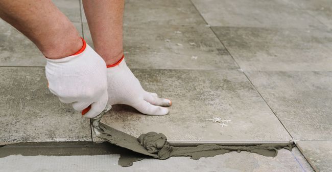 Gloved hands spreading wet mortar between gray floor tiles during repair work.