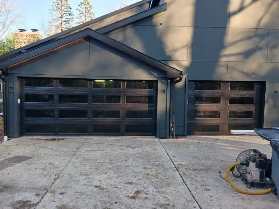 Two dark garage doors on a gray house, with a concrete driveway and a pressure washer on the right.