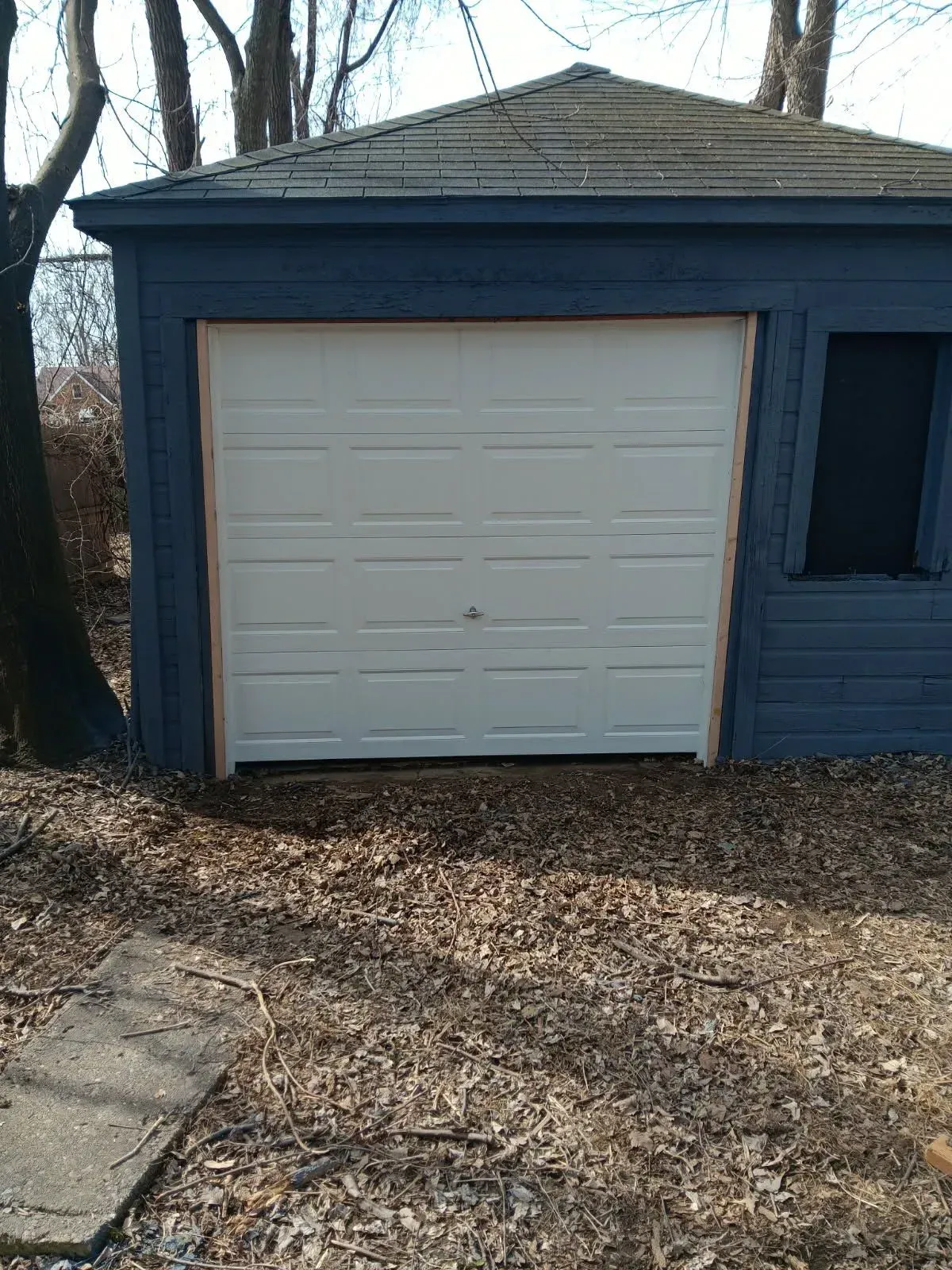 White garage door on a blue detached garage with a leaf-covered driveway and sidewalk.