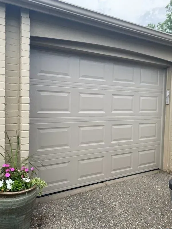 Beige garage door with paneled design beside a potted flower planter on a driveway