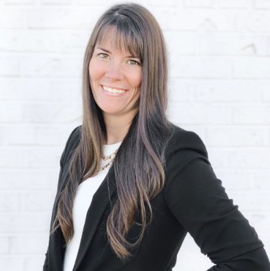 A woman in a black jacket and white shirt is smiling in front of a white brick wall.