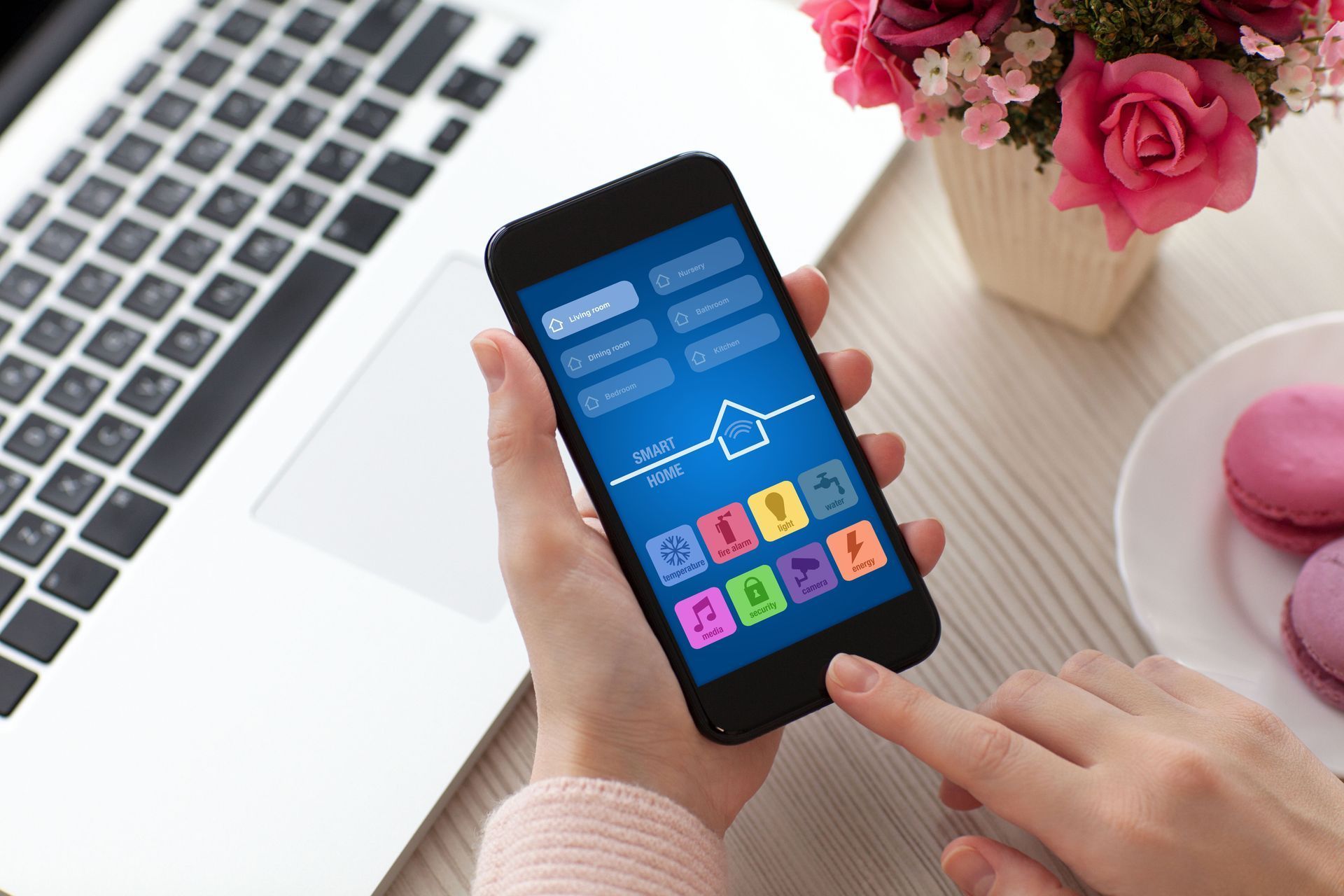 Person using a smartphone with a smart home app on a desk with a laptop and flowers.