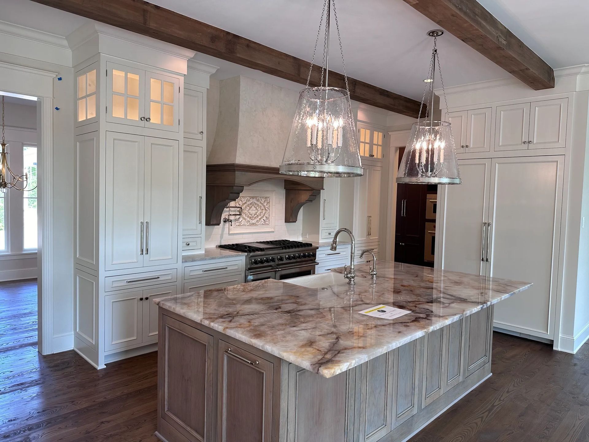 Elegant kitchen with island, white cabinetry, dark wood beams, and large chandelier lights.