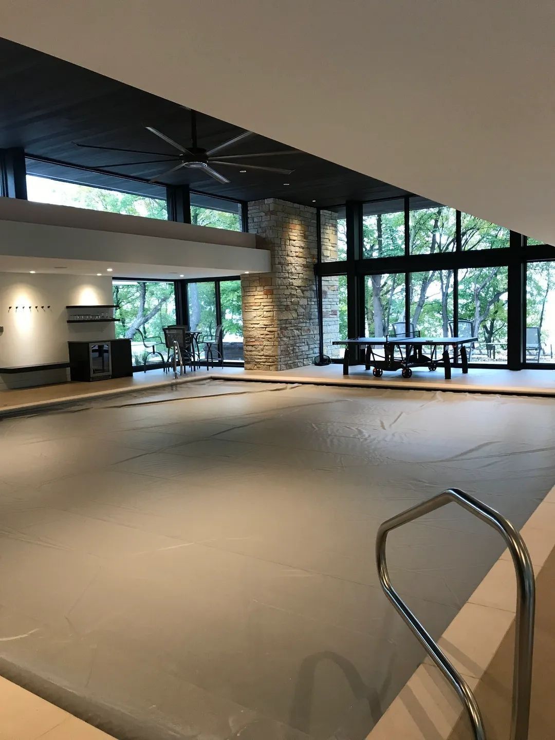 Indoor swimming pool with cover, large windows, stone column, and ceiling fan.