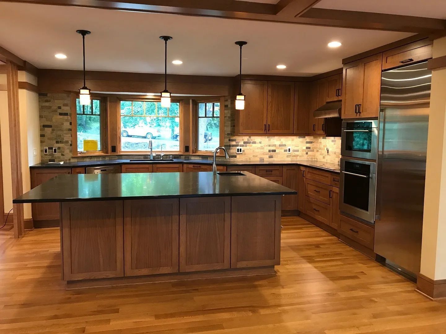 Kitchen with wood cabinets, island, and stainless steel appliances. Black countertops, stone backsplash, and hardwood floors.