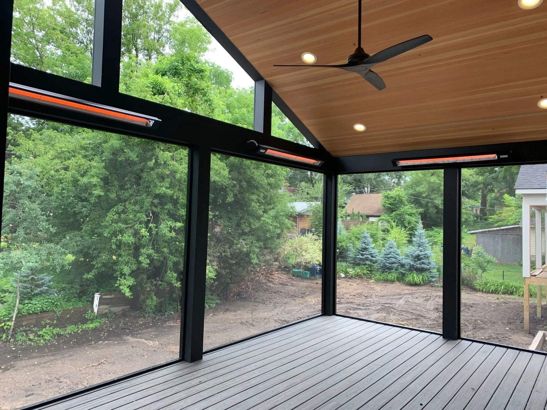 Screened-in porch with dark frame, gray floor, wood ceiling, heaters, ceiling fan, overlooking a backyard with trees.