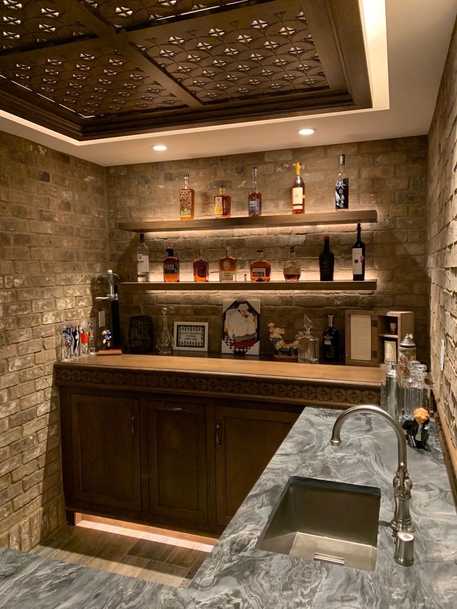 Home bar with brick walls, wood shelves displaying liquor bottles, and a stainless steel sink.