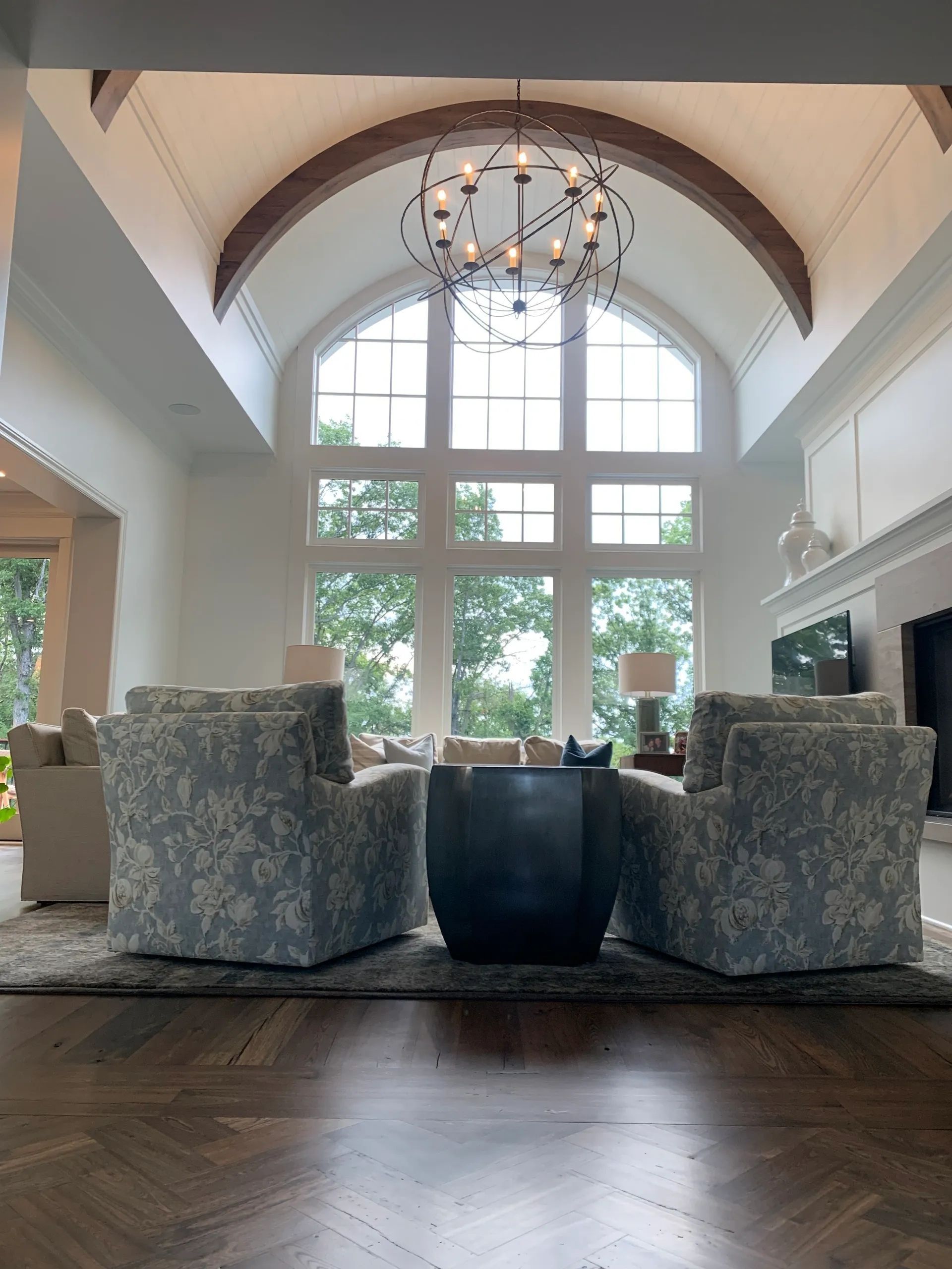 A spacious living room with large windows, patterned armchairs, and a large chandelier.