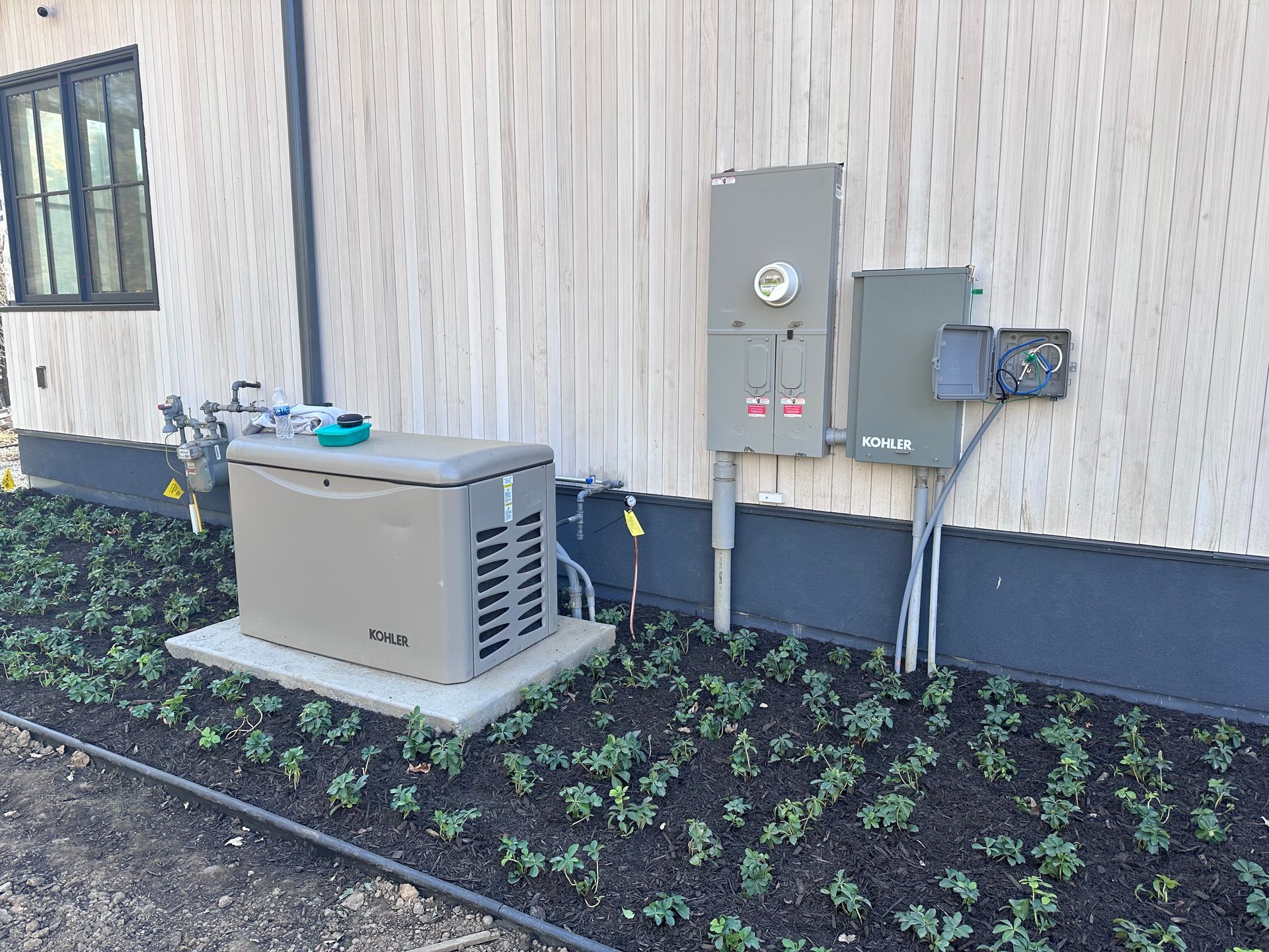 Generator and electrical boxes mounted on a house exterior next to a small garden.