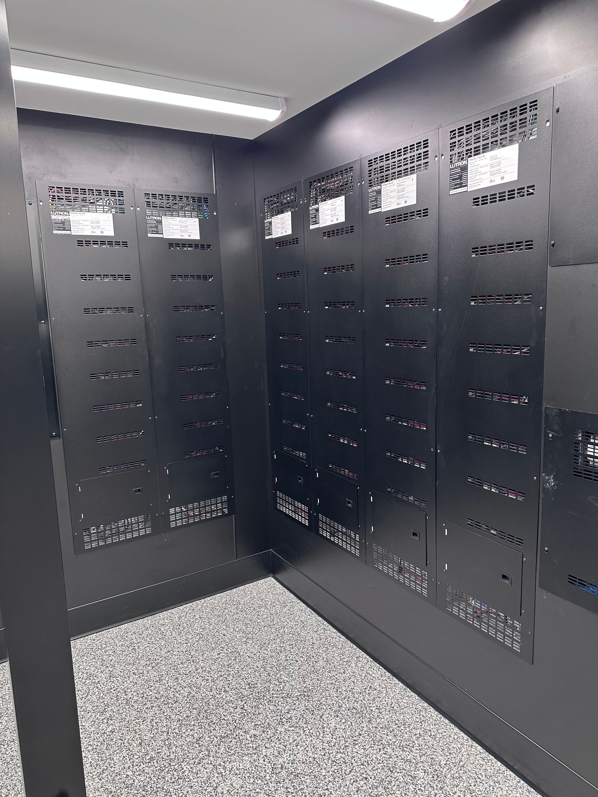 Black metal server racks in a corner, with perforated panels and a speckled floor.