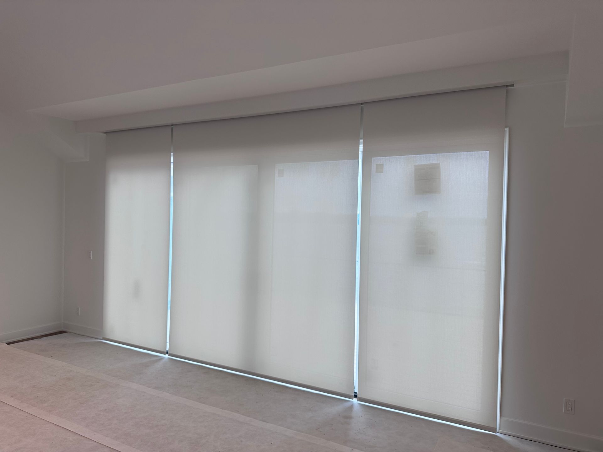 Four closed, off-white roller blinds cover a wide window in a room with light carpet and white walls.