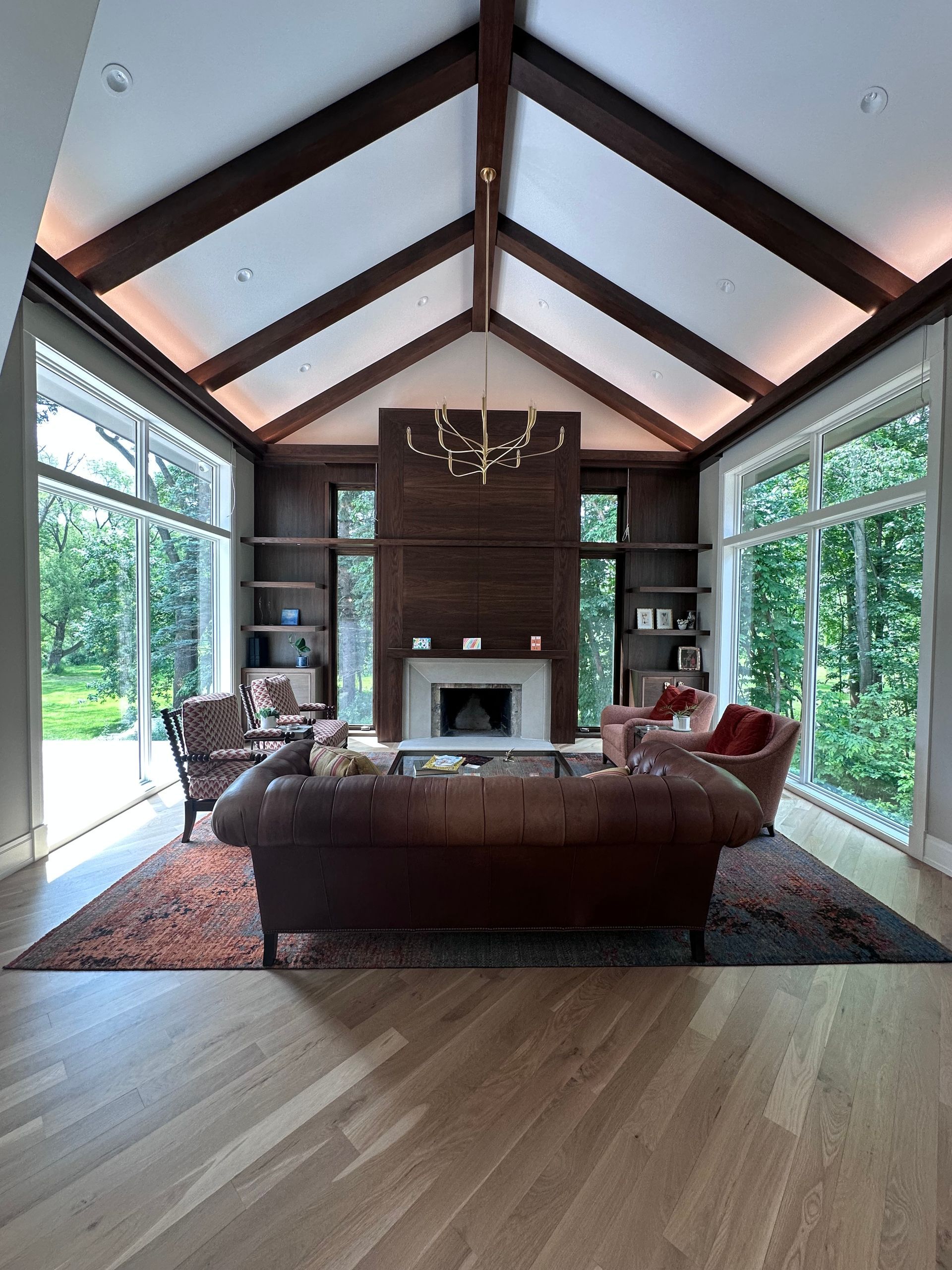 Living room with high, wood-beamed ceiling, large windows, fireplace, and furniture on a patterned rug.