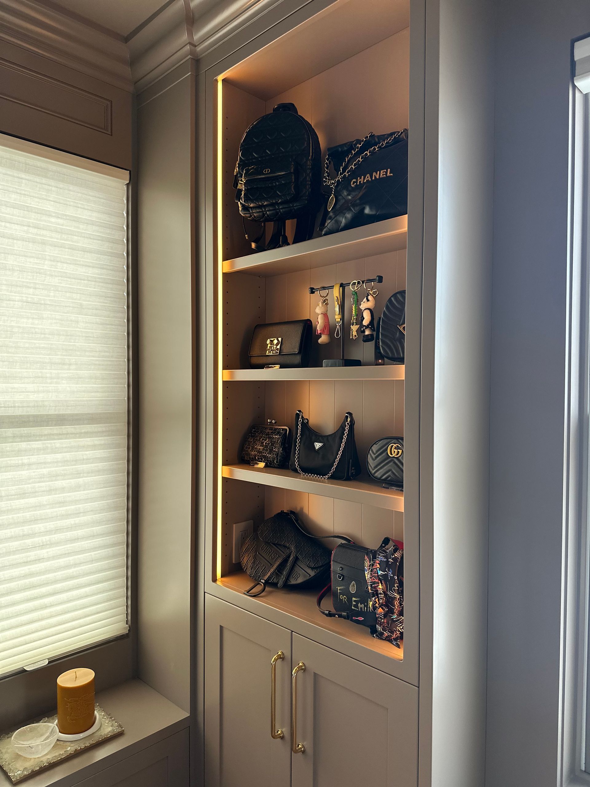 Built-in cabinet with shelves displaying handbags and a backpack. Lit with warm light, beside a window with blinds.