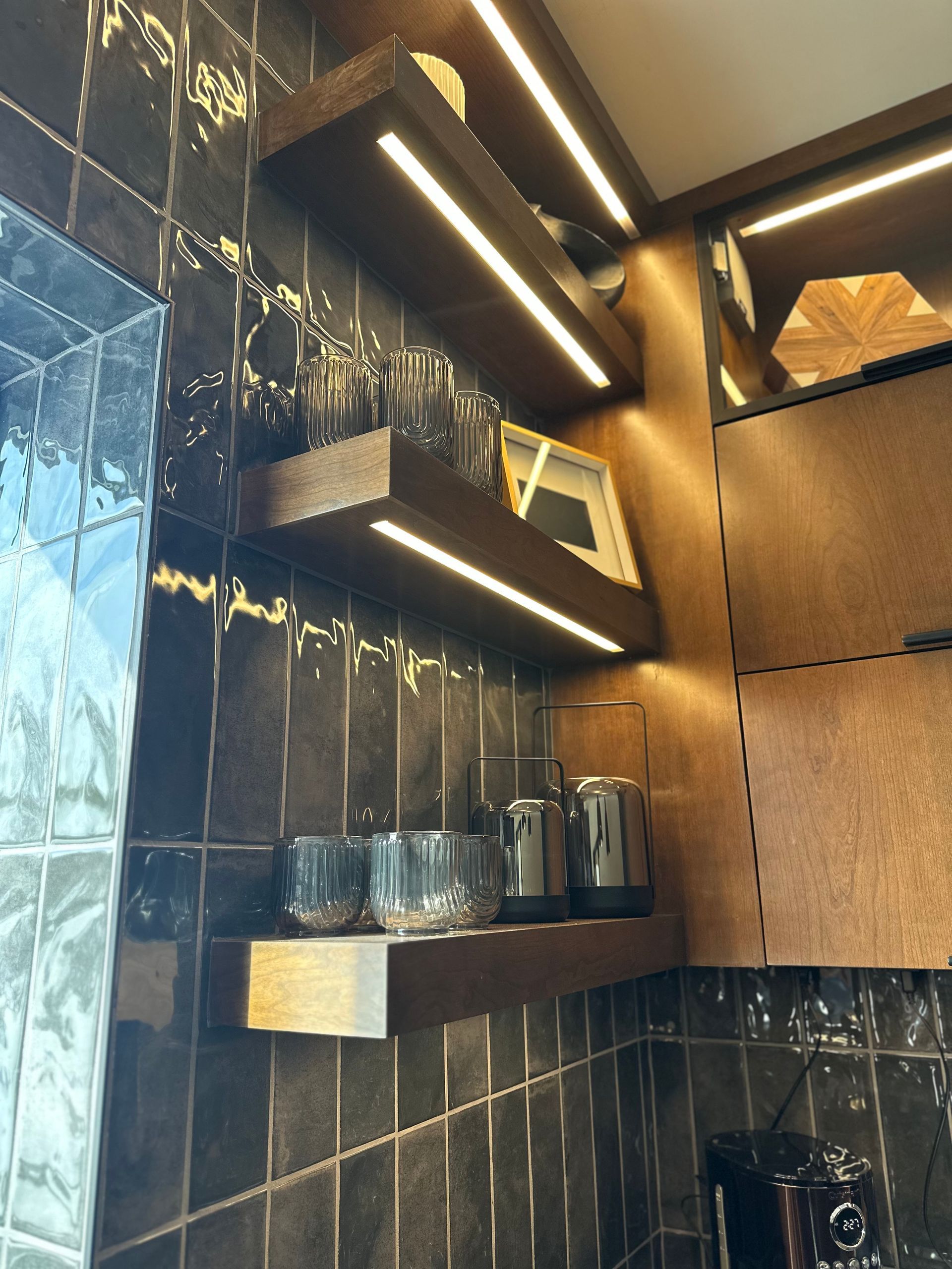 Dark wooden shelves with LED lighting in a kitchen, holding glassware and objects against a black tiled wall.