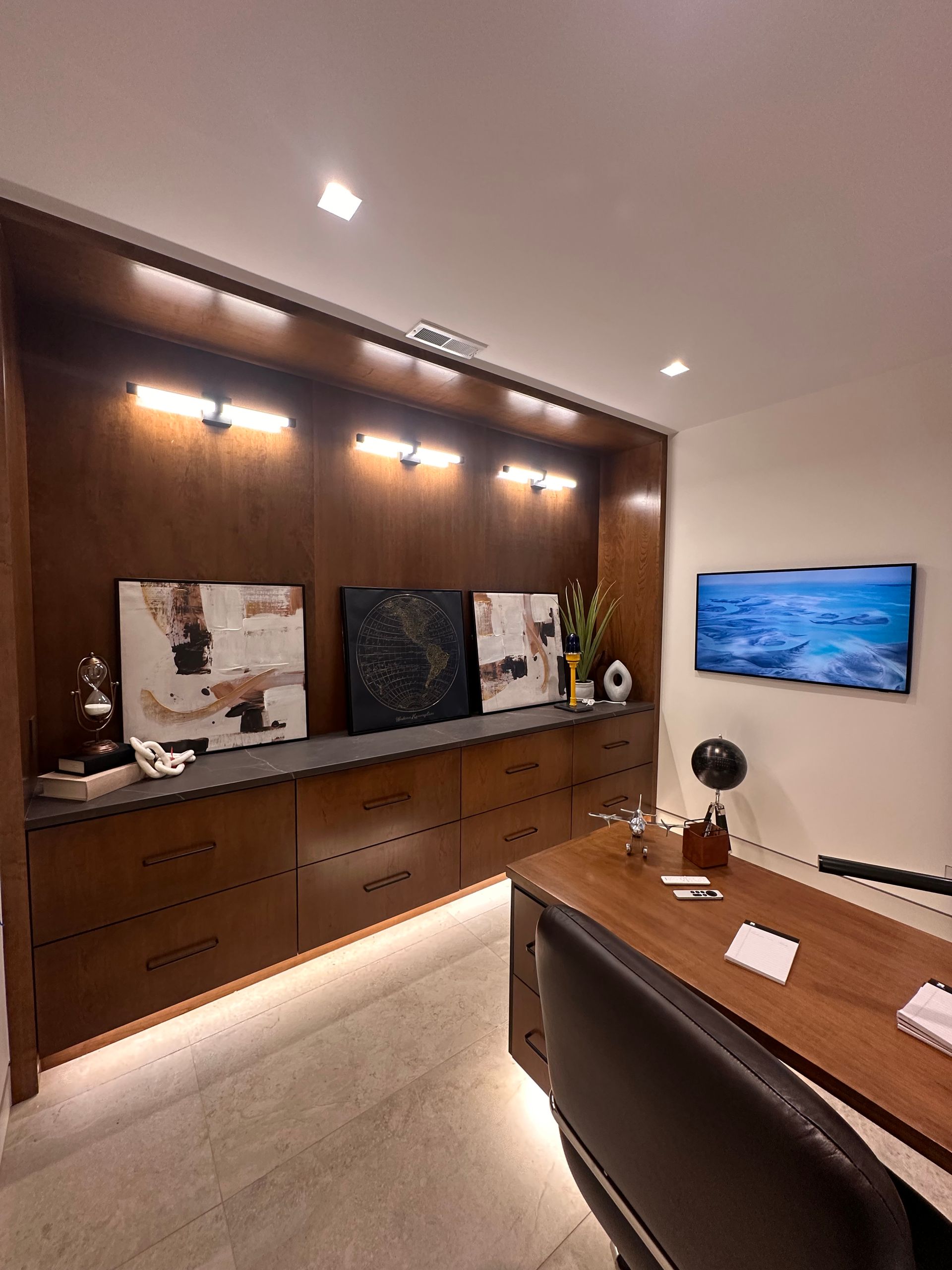 Office with built-in dark wood cabinetry, artwork, desk, and a television. Warm lighting illuminates the space.