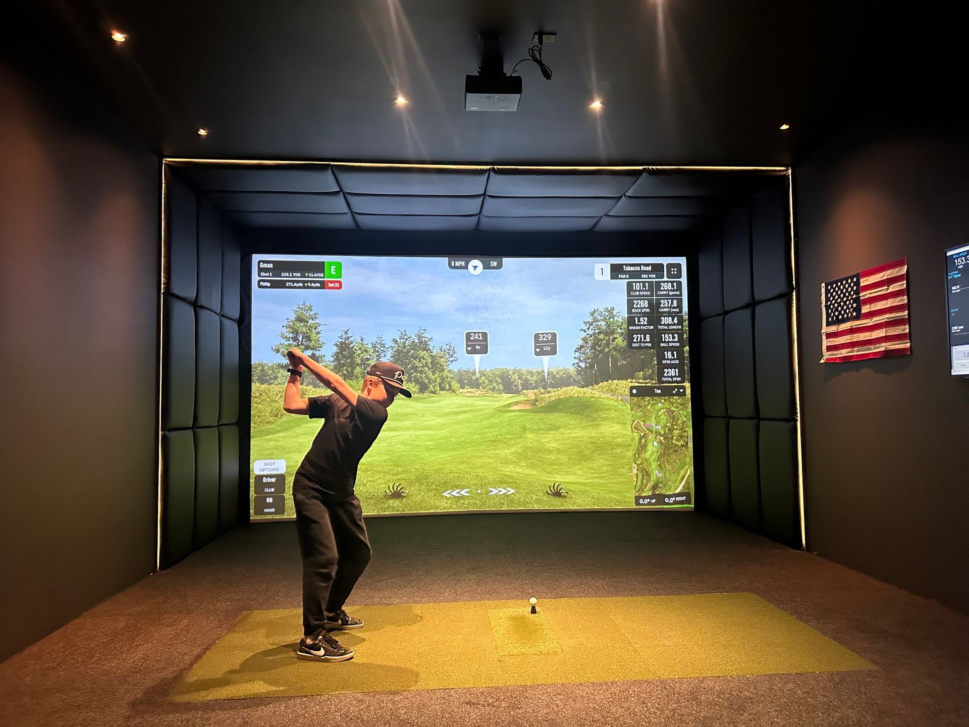 Person swings golf club in a golf simulator. Dark room, large screen showing a course, golf ball on the ground.