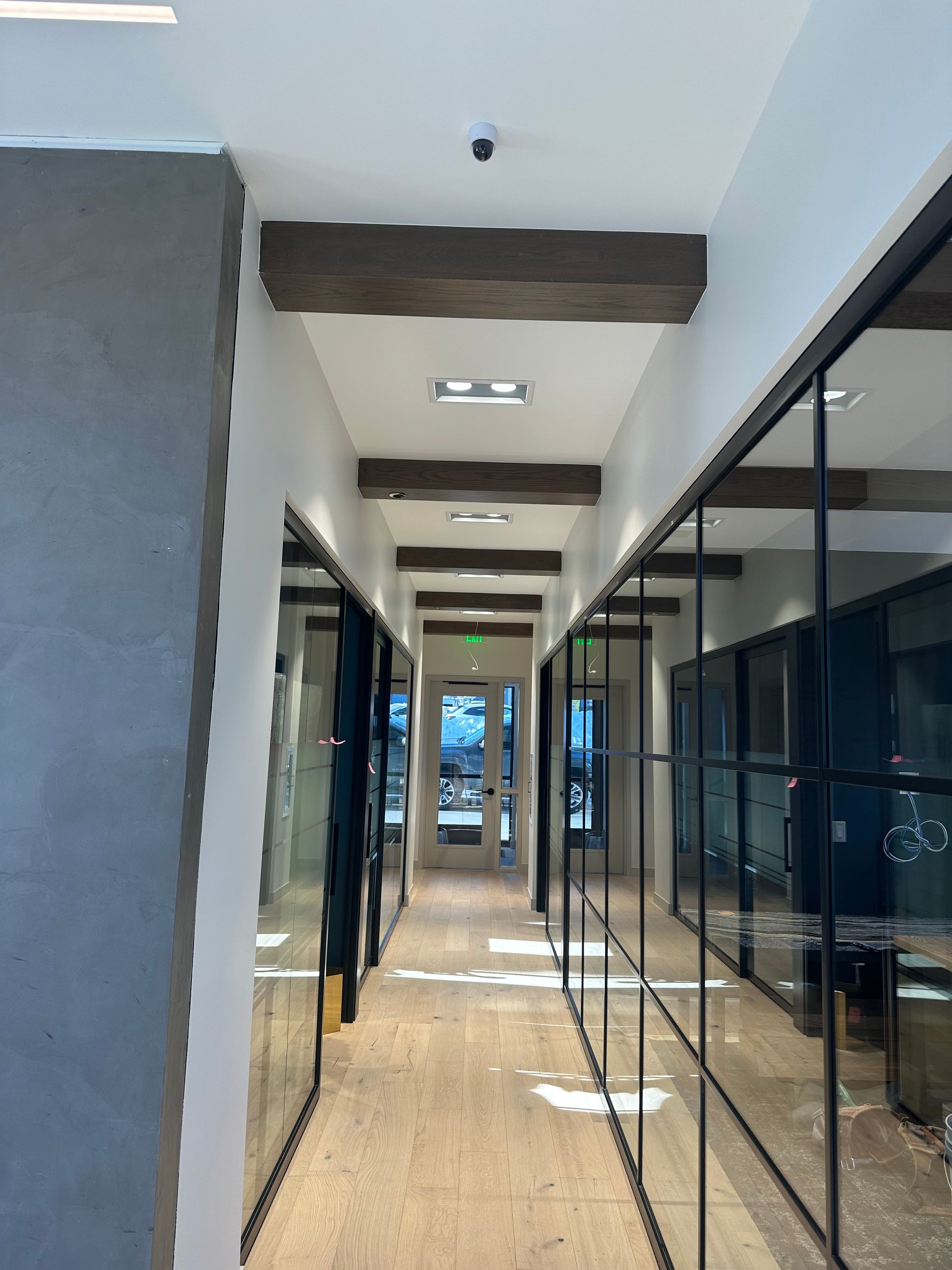 Hallway with glass-walled offices. Wooden beams and light fixtures on the ceiling. Light wood flooring.