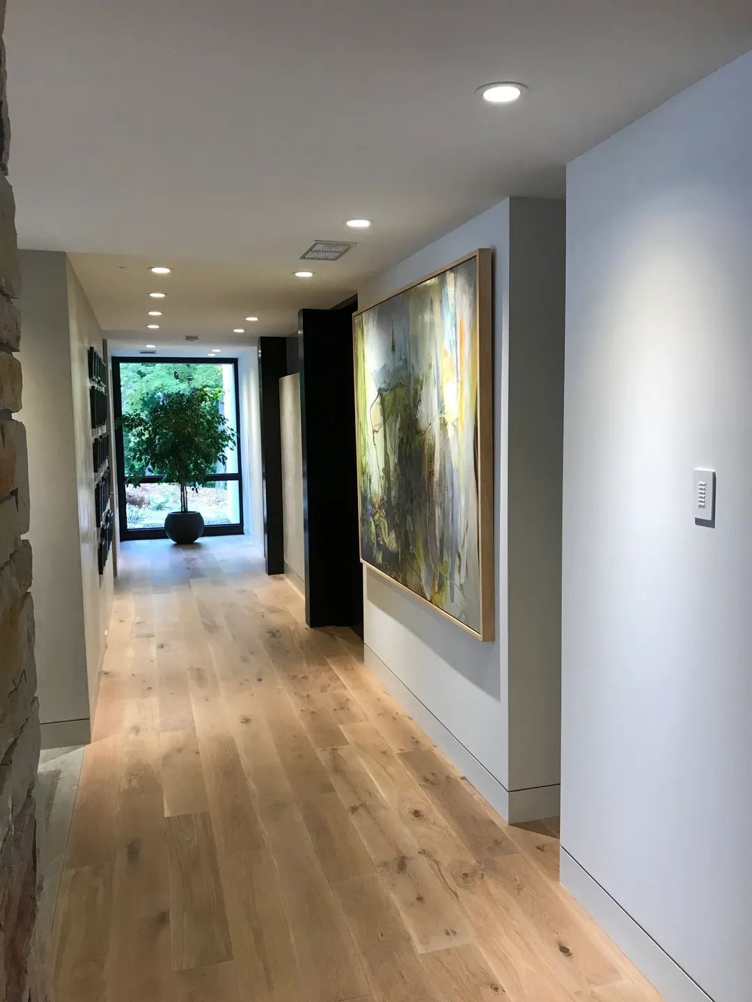 Long hallway with light wood floor, white walls, artwork, and recessed lighting.