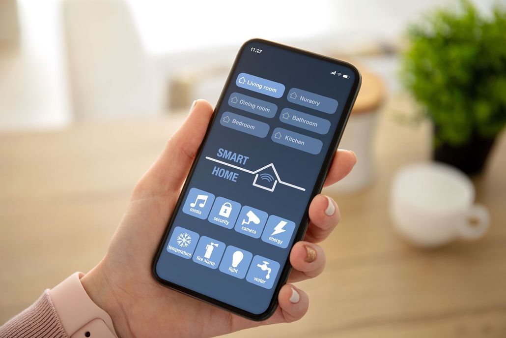 A hand holds a smartphone displaying a smart home control app with various icons for managing home devices.