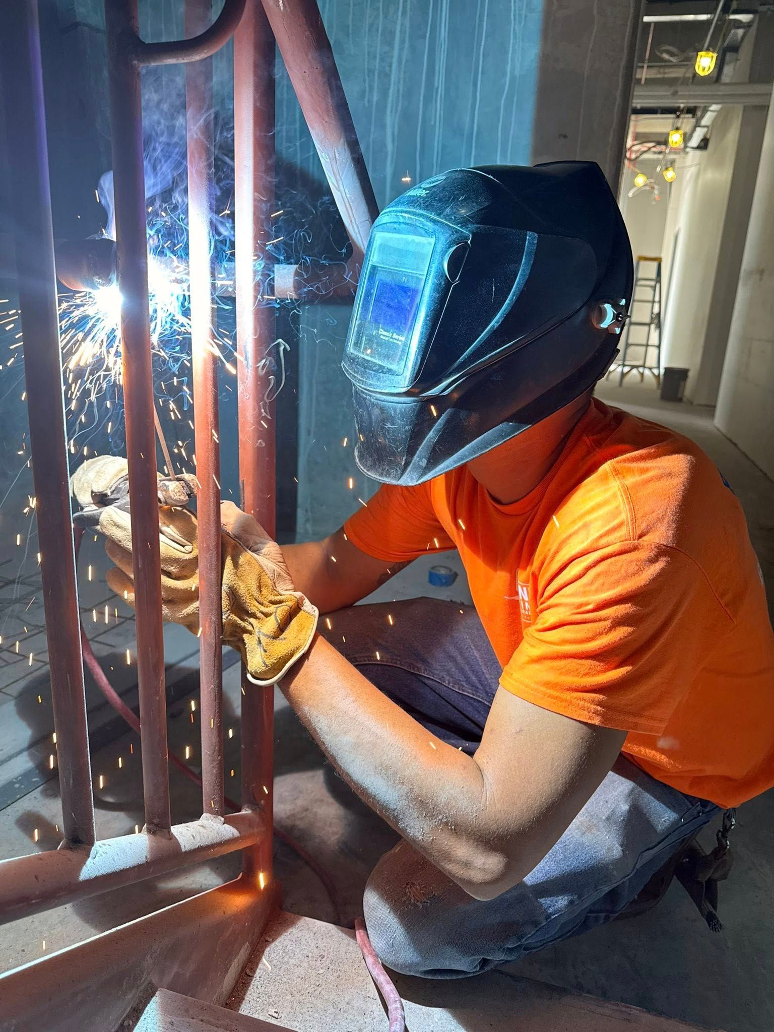 A person wearing a welding helmet and an orange shirt arc welds a metal railing indoors, surrounded by sparks.