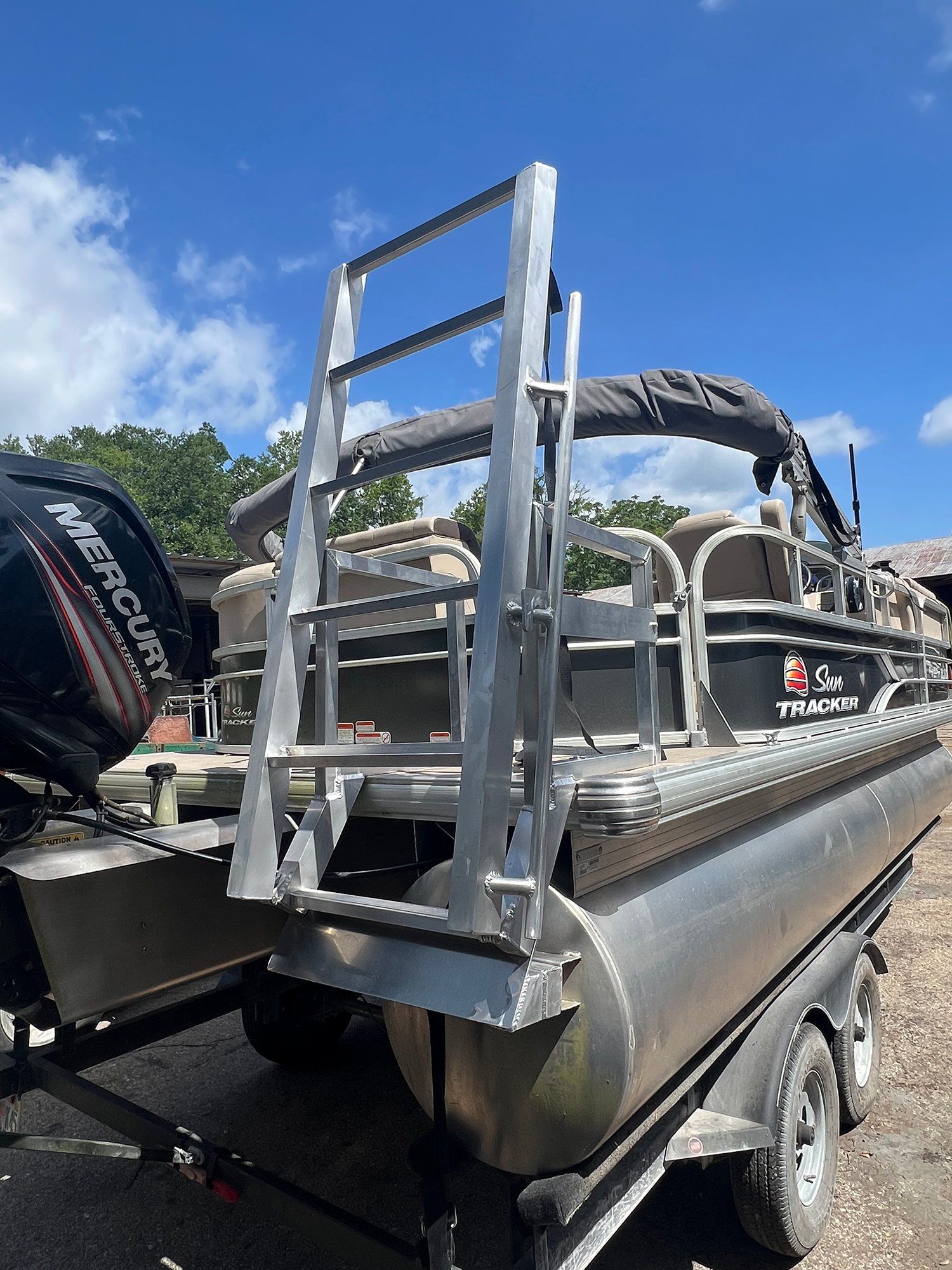 A silver metal ladder attached to the rear of a pontoon boat on a trailer outdoors under a clear blue sky.
